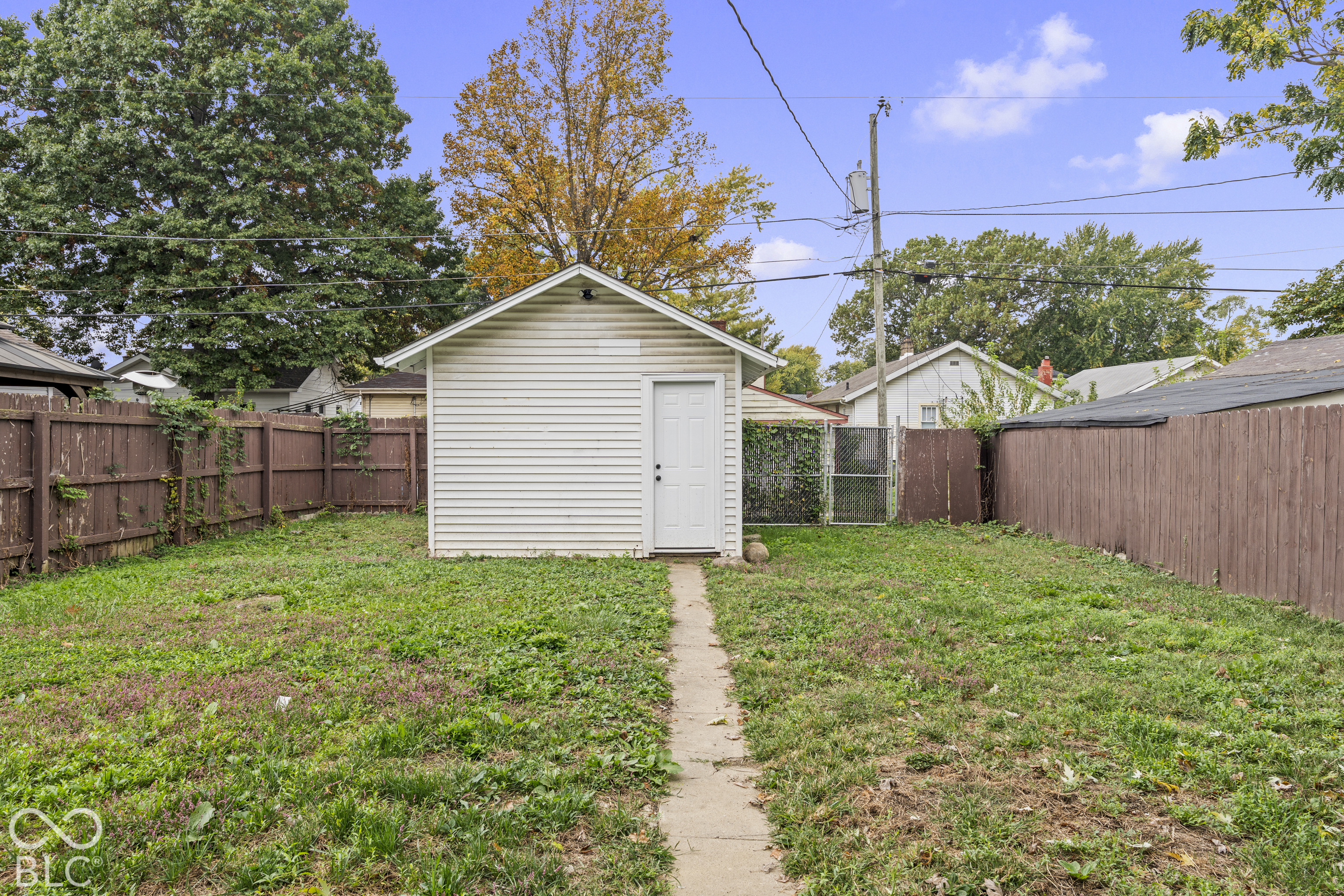 Detached exterior structure with white horizontal siding and a single entry door, situated on a lot enclosed by wood panel fencing, featuring a concrete pathway Detached exterior structure with white horizontal siding and a single entry door, situated on a lot enclosed by wood panel fencing, featuring a concrete pathway