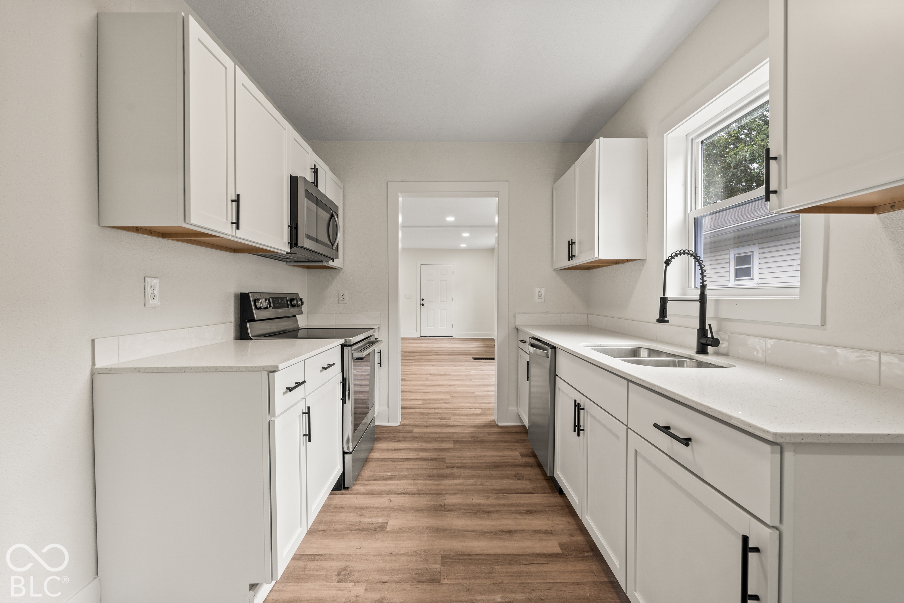 Kitchen featuring white shaker-style cabinetry, solid-surface countertops, wood-finish flooring, stainless steel appliances, and a matte black gooseneck faucet Kitchen featuring white shaker-style cabinetry, solid-surface countertops, wood-finish flooring, stainless steel appliances, and a matte black gooseneck faucet