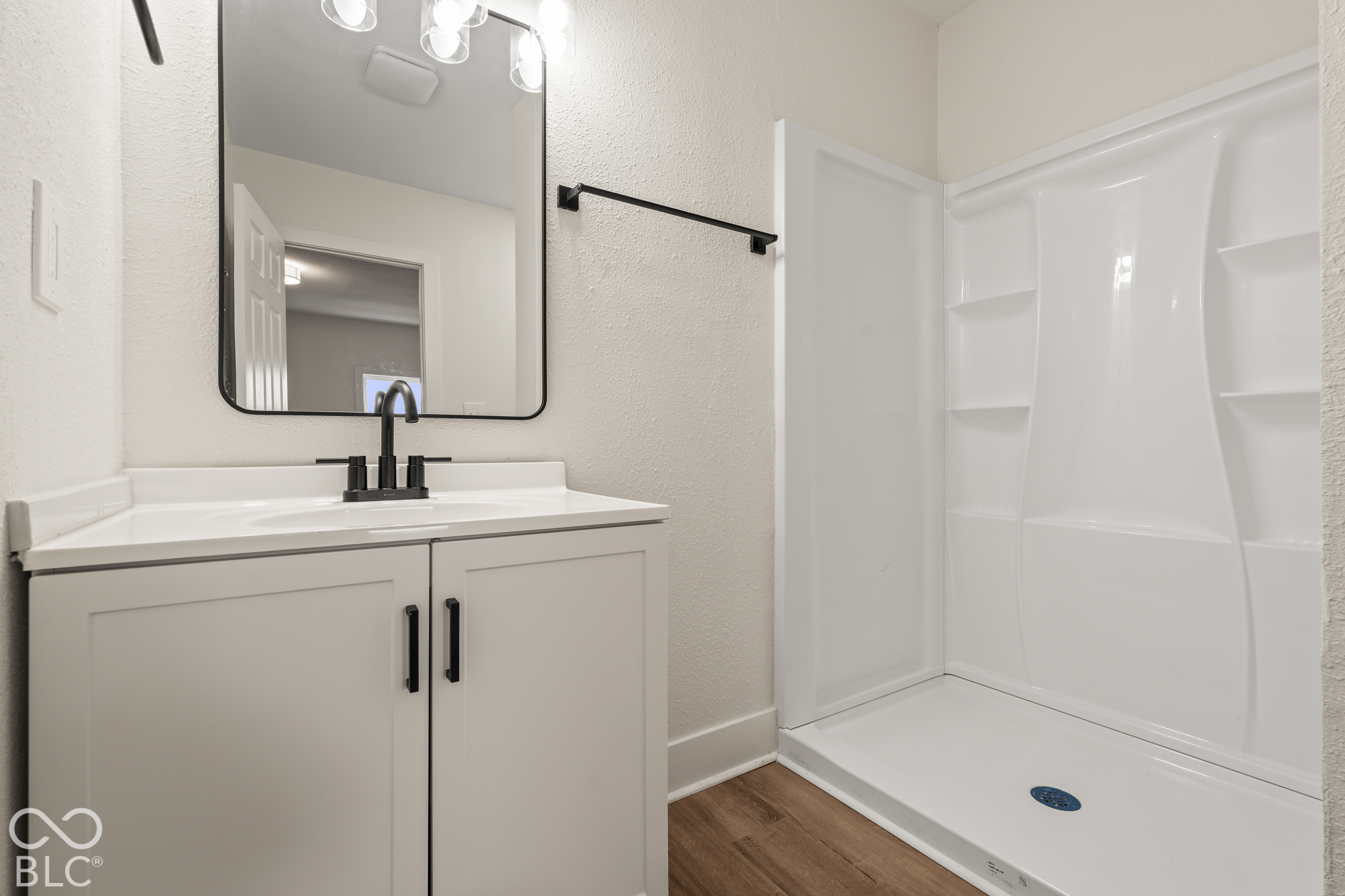 Bathroom featuring a vanity with white cabinetry, a rectangular mirror with a rounded black frame, a matte black faucet, and a white walk-in shower surround