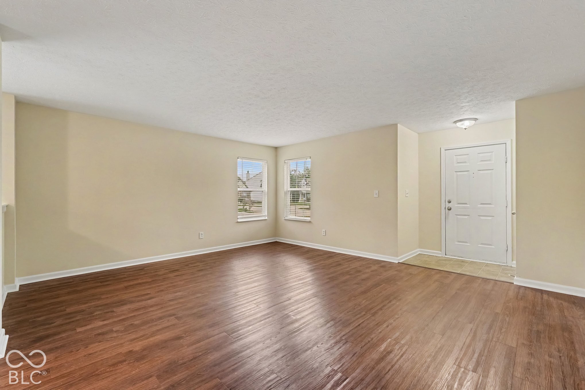 Open-concept interior featuring wood-finish flooring, neutral wall paint, dual windows, and an exterior entry door with a flush-mount light fixture