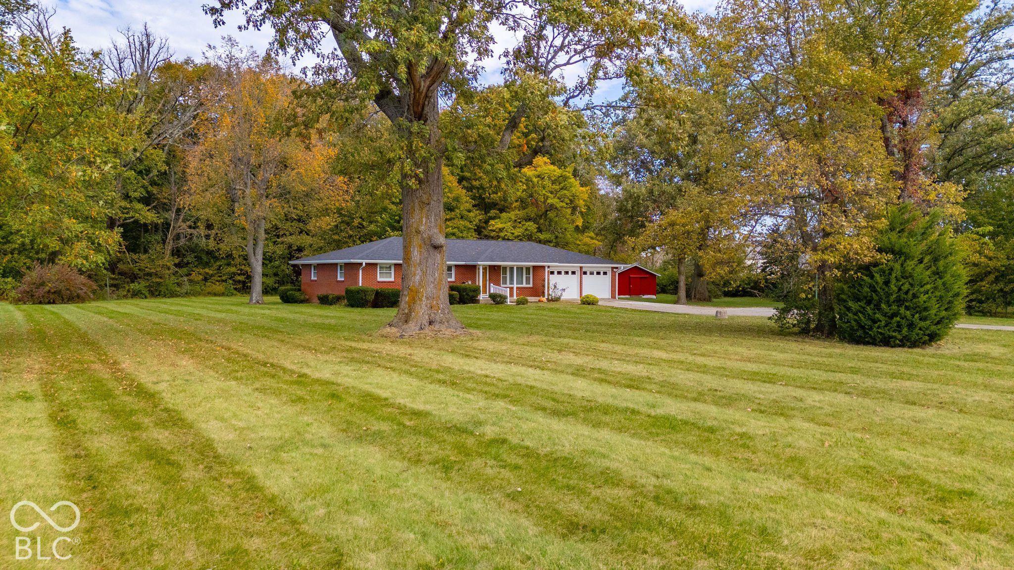 Single-story brick residence featuring an attached two-car garage, asphalt shingle roof, expansive manicured lawn, and mature trees on a wooded lot