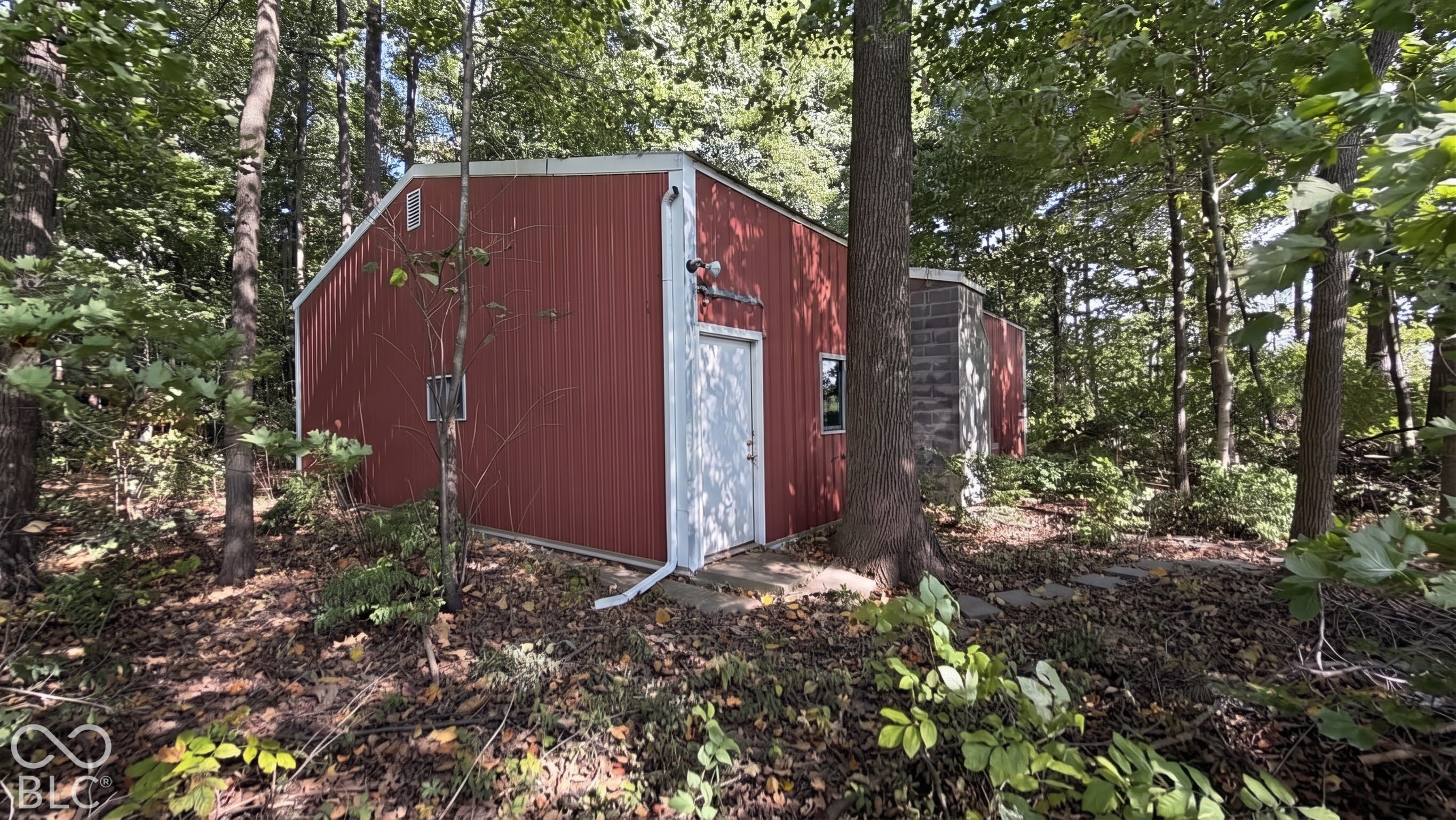 Detached structure with vertical metal siding, masonry chimney, and metal door, situated in a wooded setting