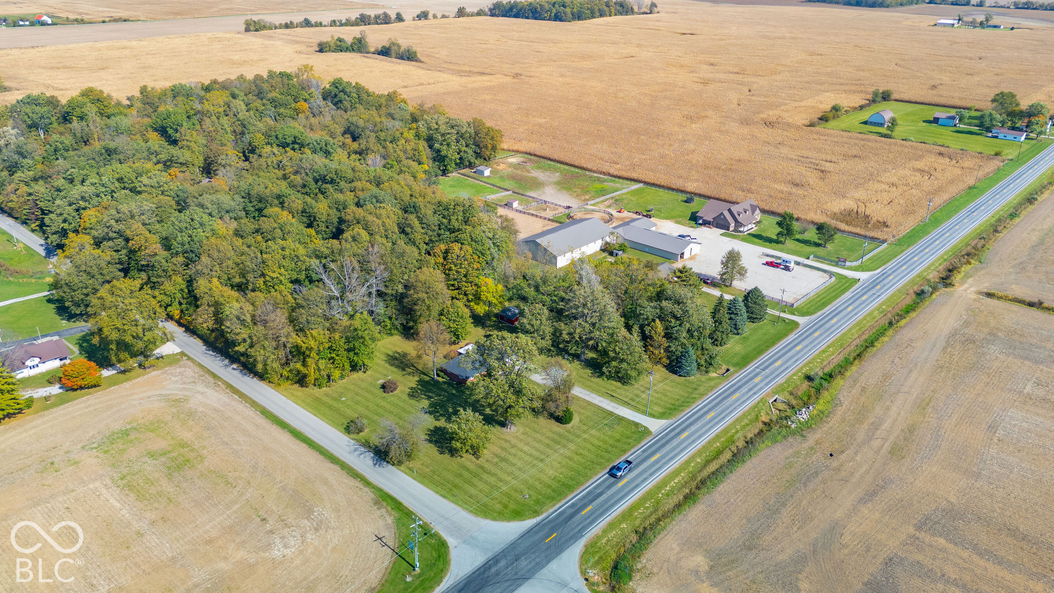 Aerial perspective of property featuring extensive wooded acreage, open lawn space, multiple outbuildings, and paved road access