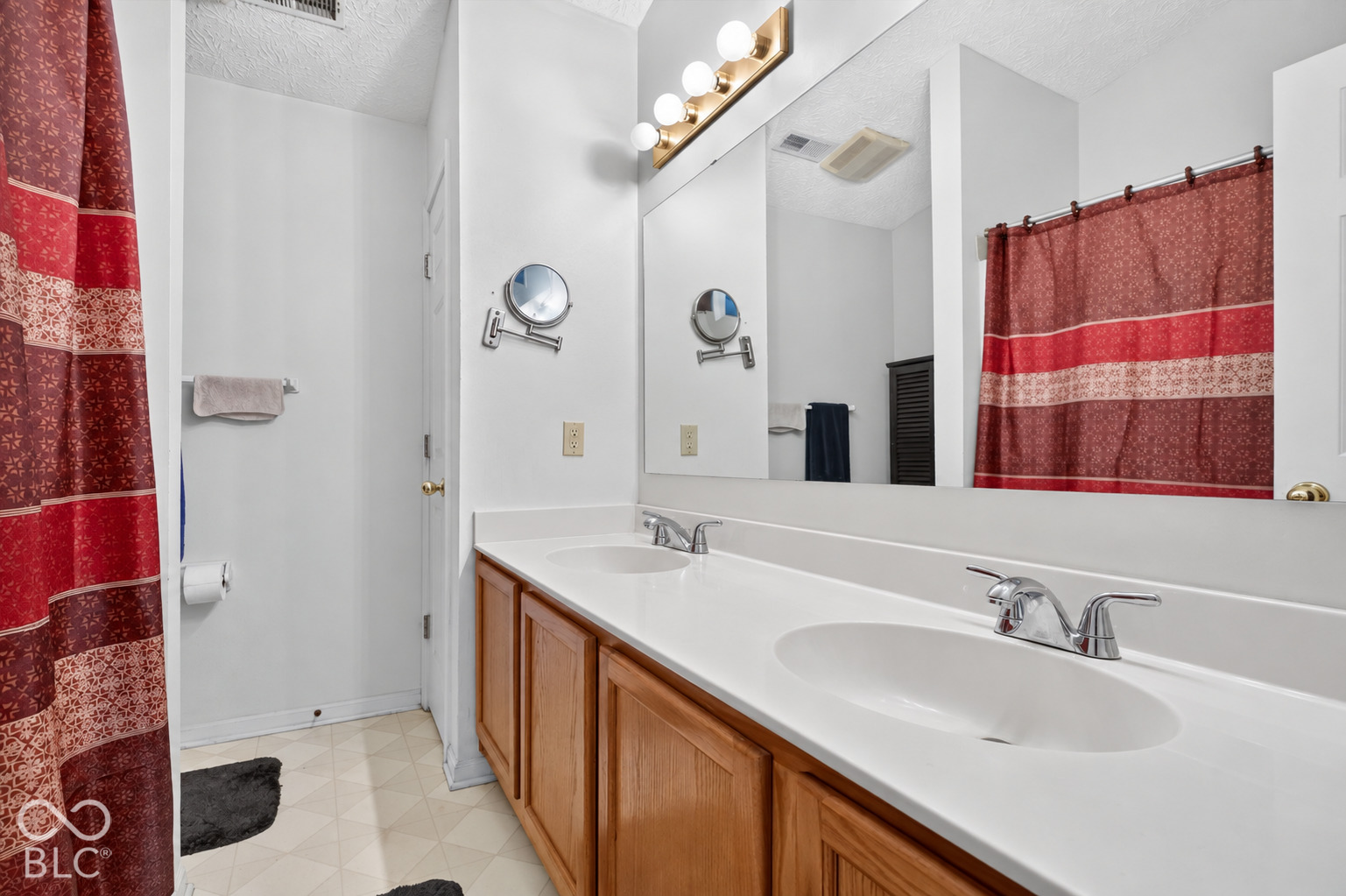 Bathroom vanity featuring a dual-basin countertop, wood-finish cabinetry, a full-width wall mirror, and a multi-bulb light fixture