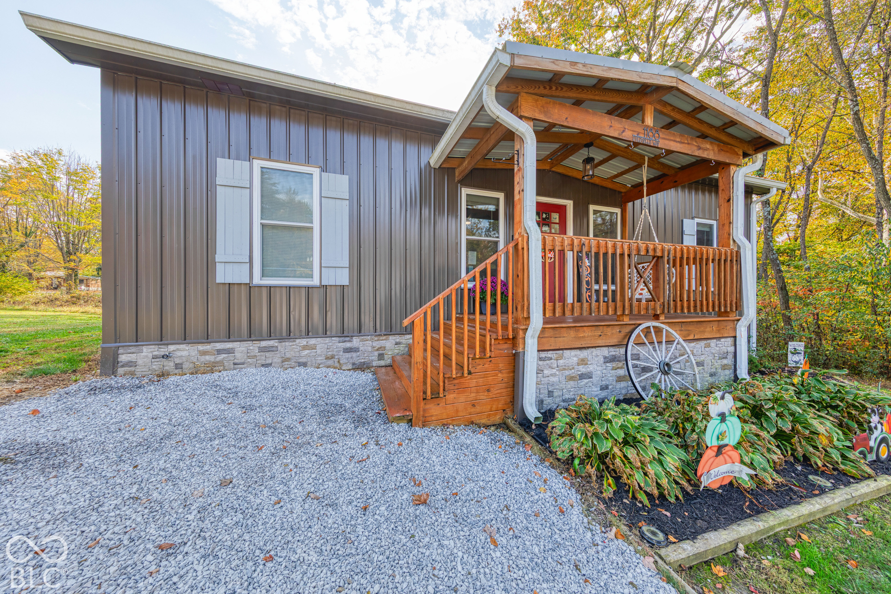 Metal-sided exterior with stone veneer skirting, covered front porch featuring a wooden railing, and a gravel driveway
