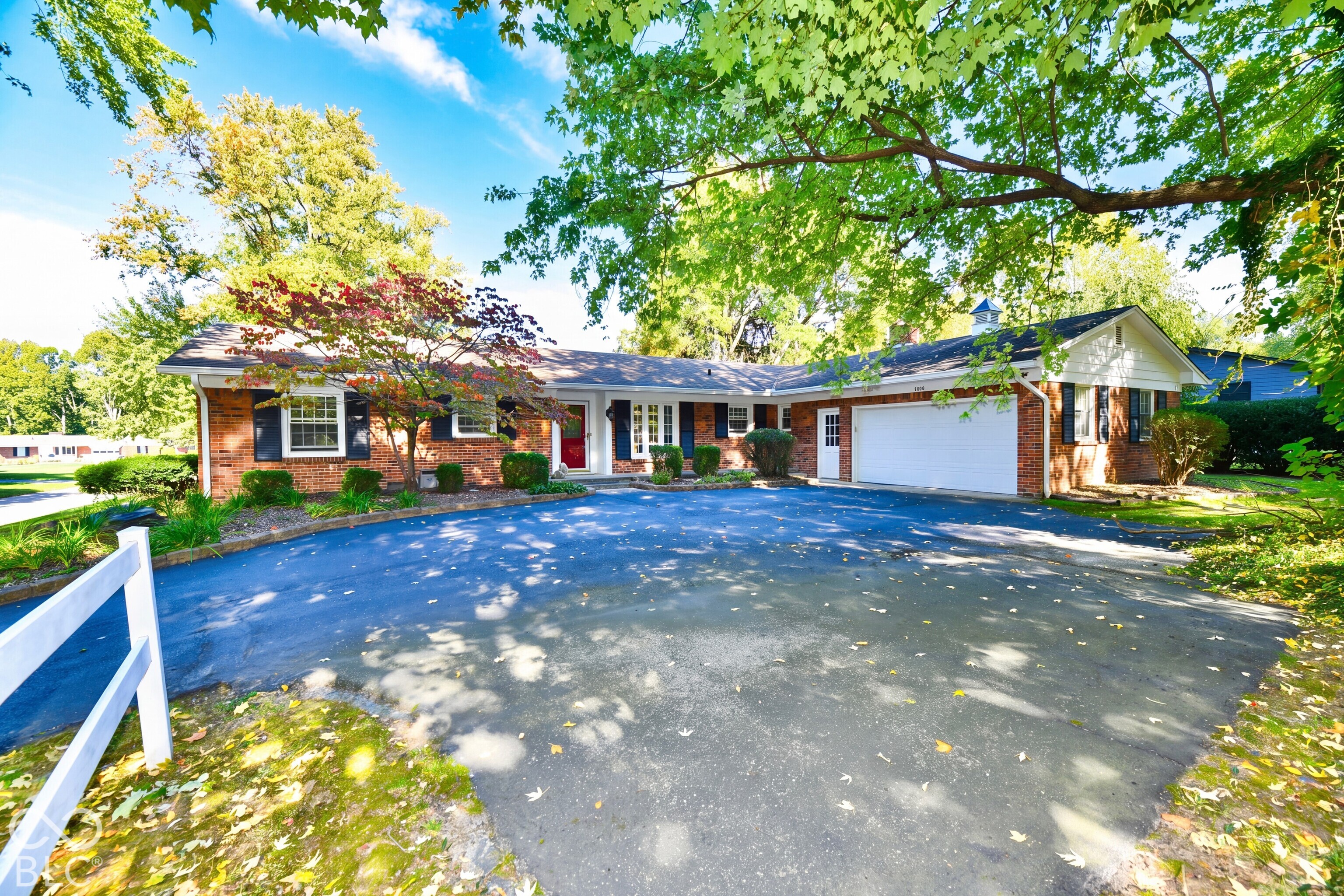 Single-story brick exterior featuring a white attached two-car garage, dark decorative shutters, and an expansive asphalt driveway Single-story brick exterior featuring a white attached two-car garage, dark decorative shutters, and an expansive asphalt driveway