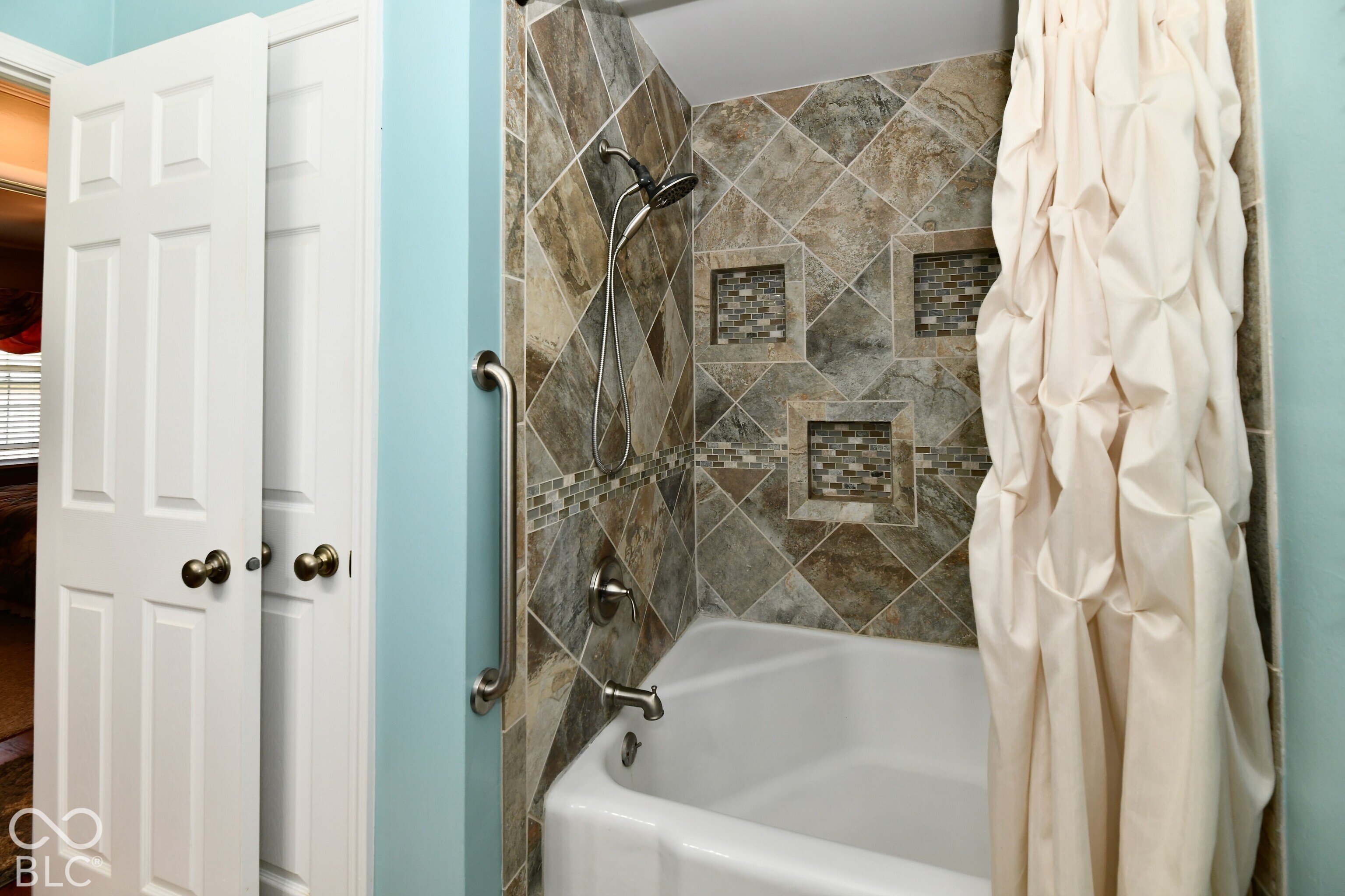 Bathtub and shower combination featuring diagonal stone-finish tile surround, integrated recessed shelving, metal grab bar, and adjustable handheld shower head
