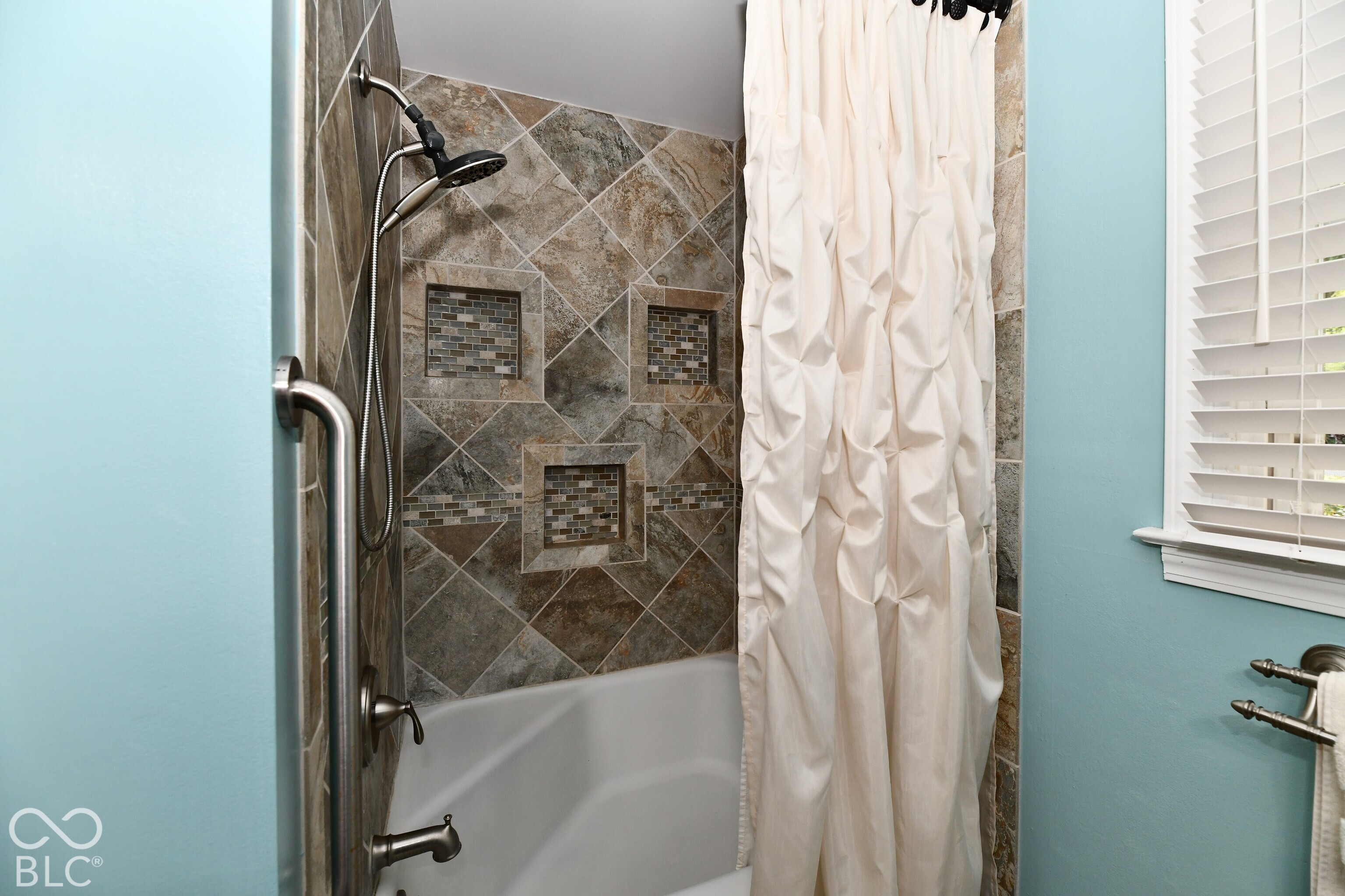 Shower featuring diagonal-set stone wall tile, built-in recessed niches, a multi-setting hand-held showerhead, metal safety grab bar, and window with adjustable blinds