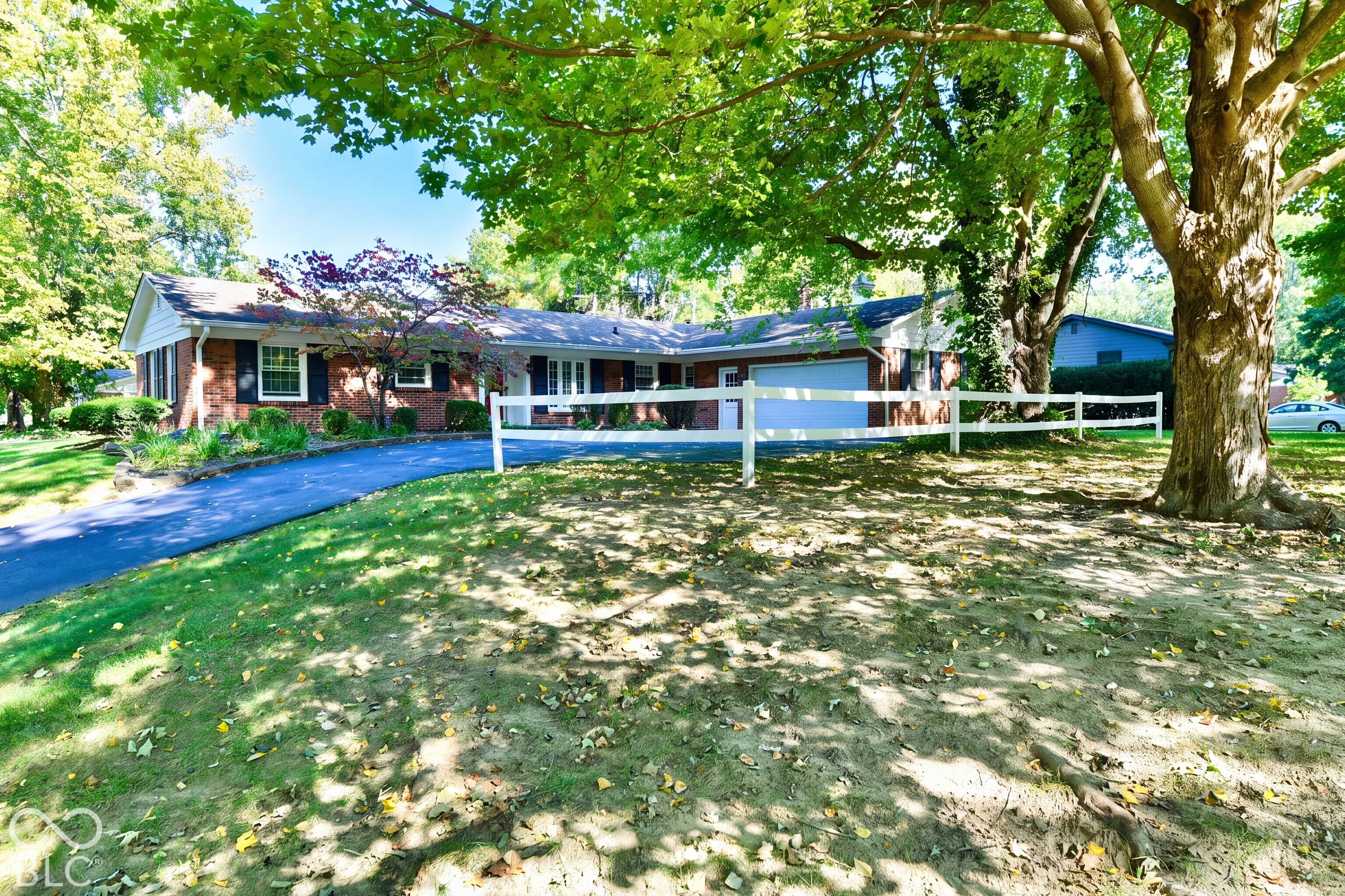 Single-story brick exterior featuring a multi-paned window facade, an attached garage, a paved driveway, and white split-rail fencing situated among mature trees