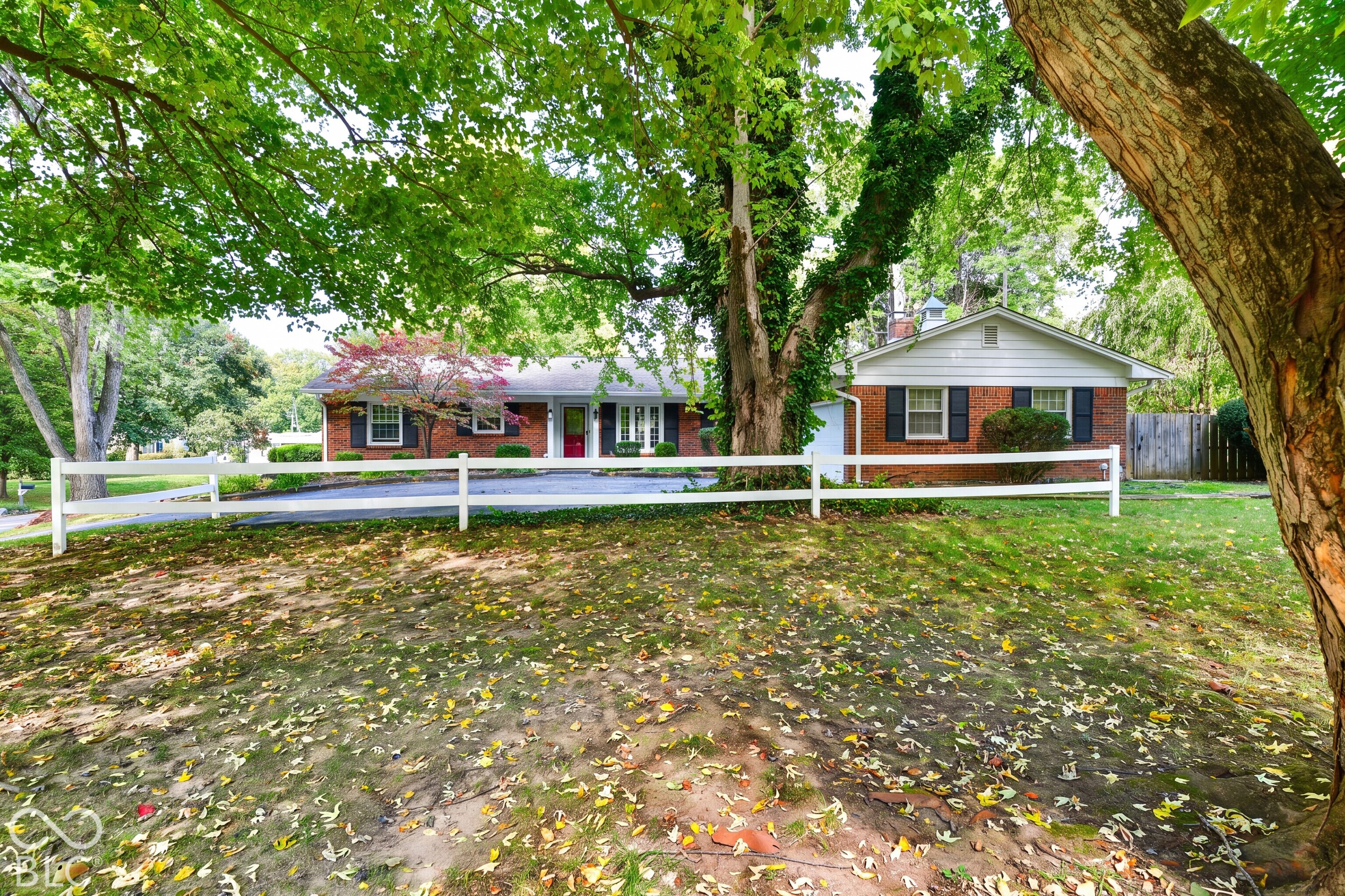 Single-story brick exterior featuring a white split-rail fence, mature trees, and shuttered windows