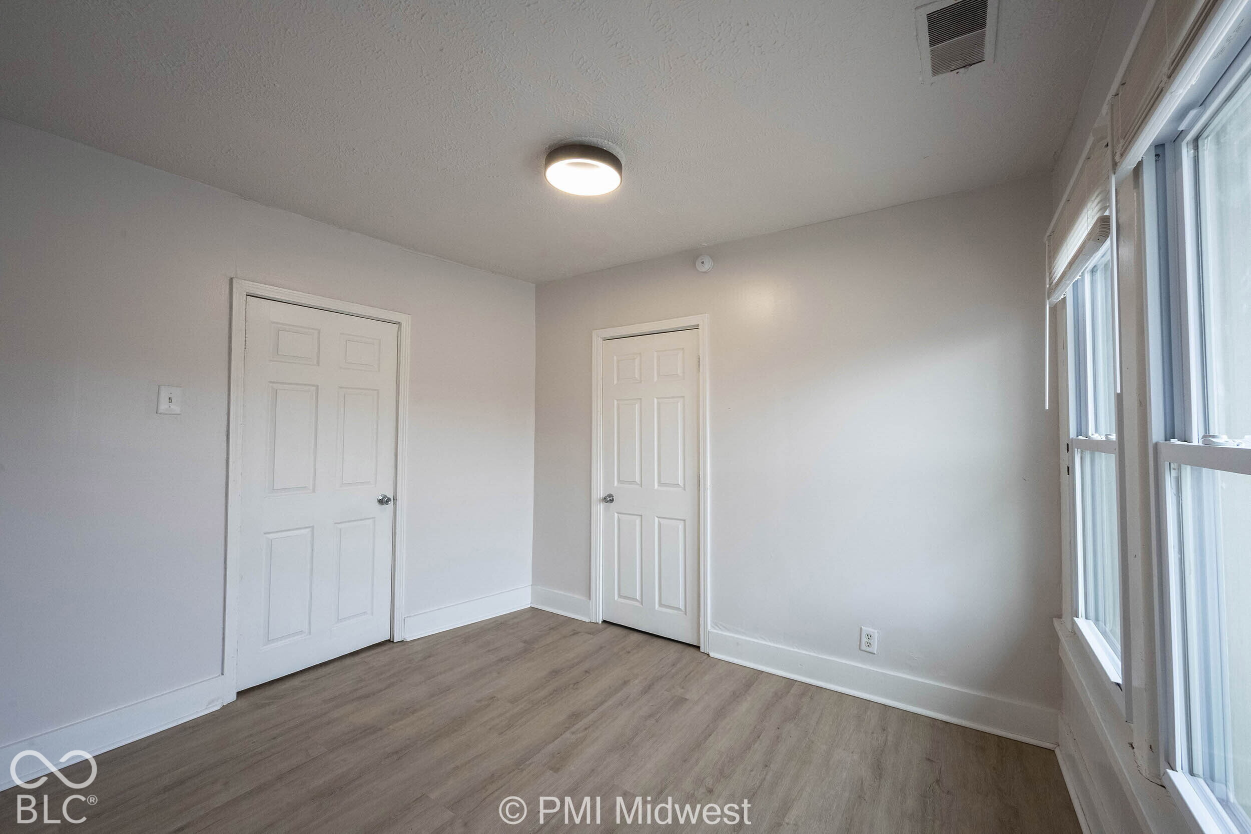 Neutral-toned interior space featuring wood-finish flooring, two six-panel doors, double-paned windows, and a flush-mount ceiling light fixture