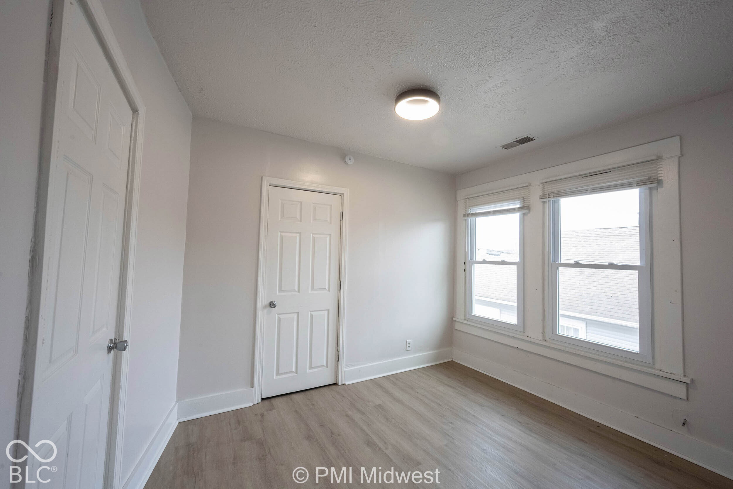 Room featuring wood-finish flooring, dual windows with white casings, two interior doors, and a flush-mount overhead light fixture