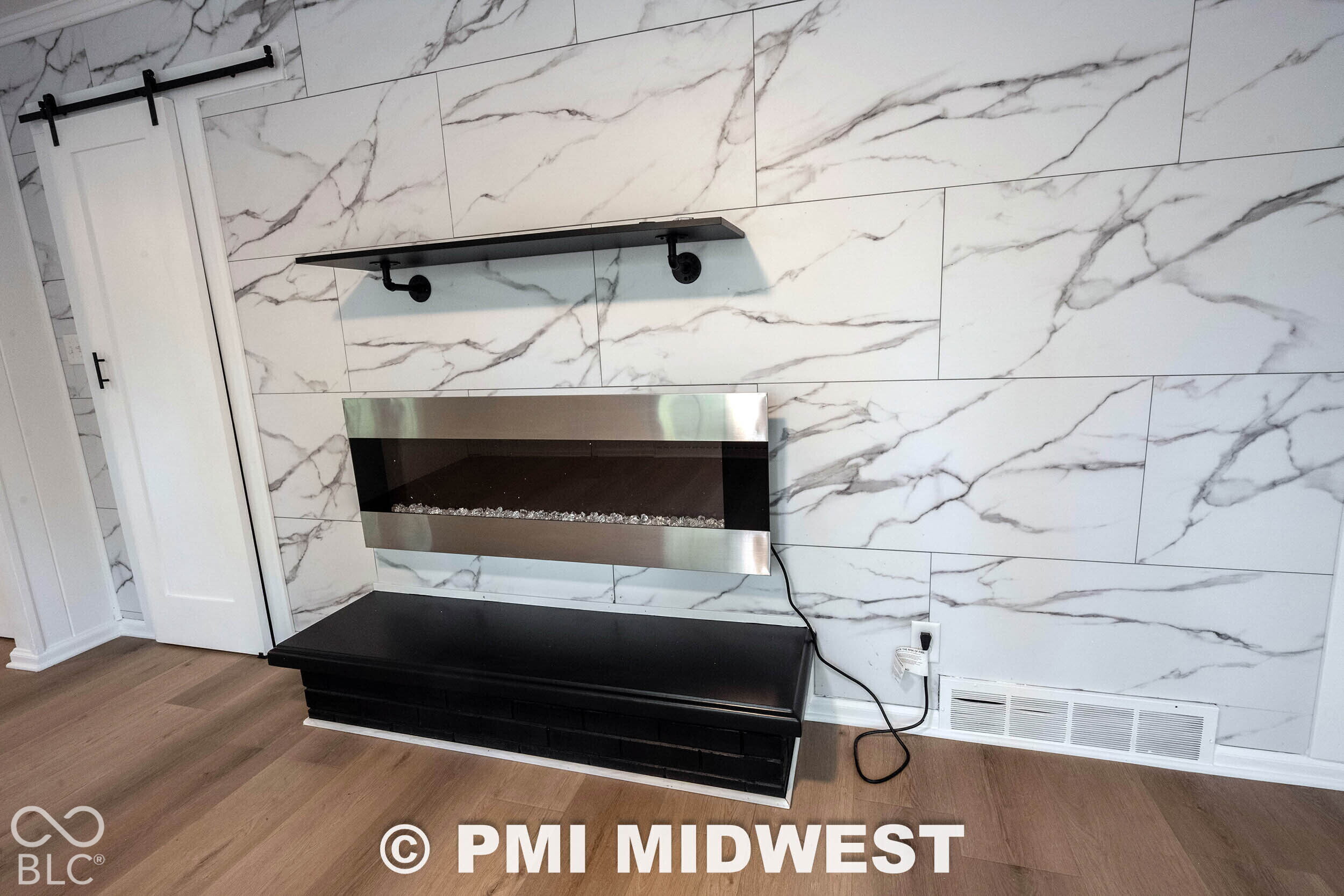 Wall-to-ceiling stone-look tile fireplace surround featuring a stainless steel wall-mounted electric fireplace, a black built-in hearth, a floating matte black shelf, and a sliding barn-style door