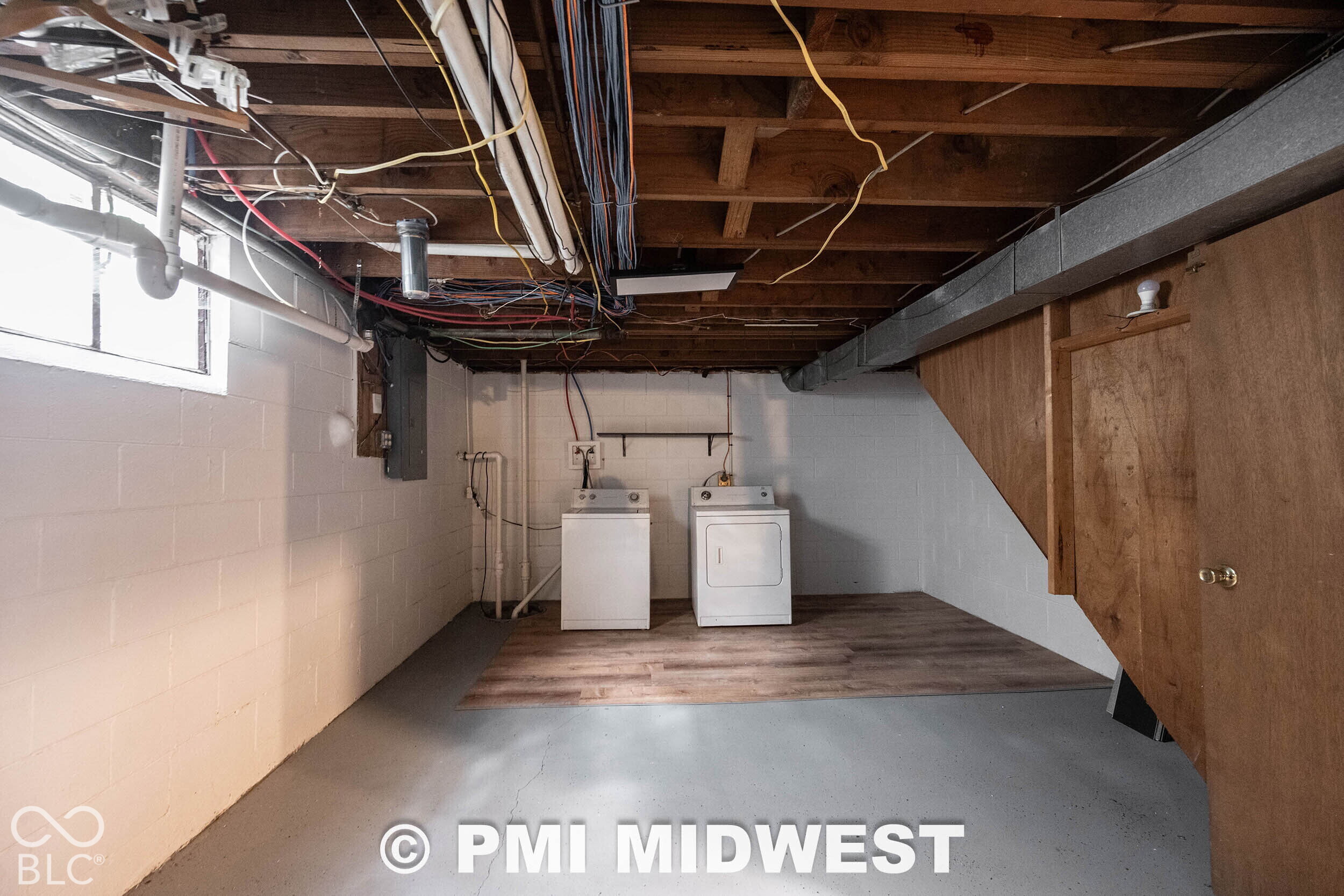 Unfinished utility area featuring exposed joists, painted concrete block walls, painted concrete floor, wood-finish flooring section, and built-in wood-panel door