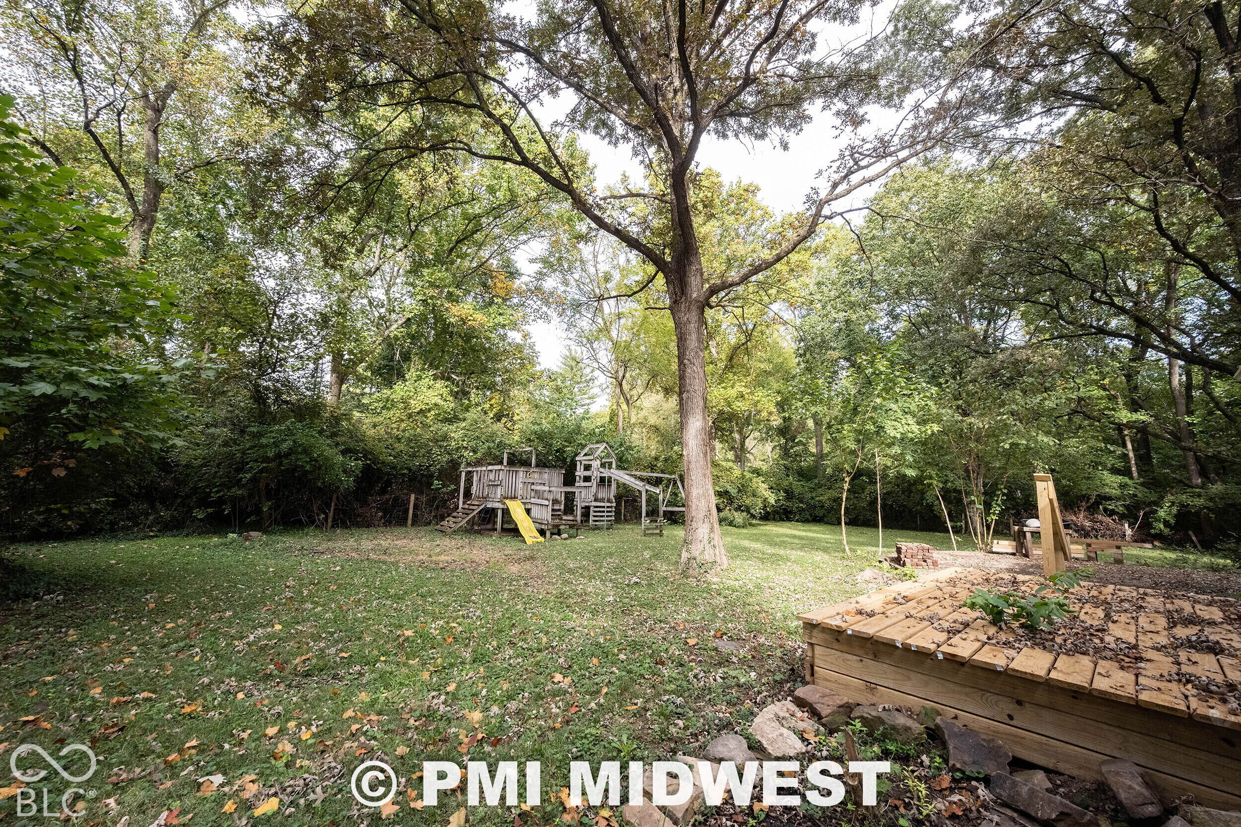 Expansive backyard featuring a leveled lawn, mature trees, tiered wood-finish decking, and stone retaining wall elements