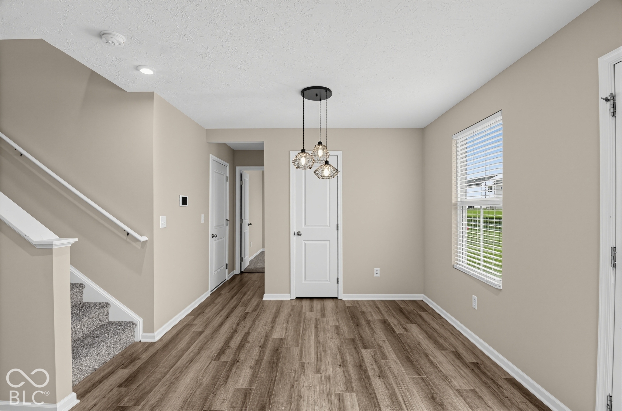 Interior space featuring wood-finish flooring, a multi-pendant light fixture, a staircase with a white handrail, neutral-toned walls, and a window with horizontal blinds