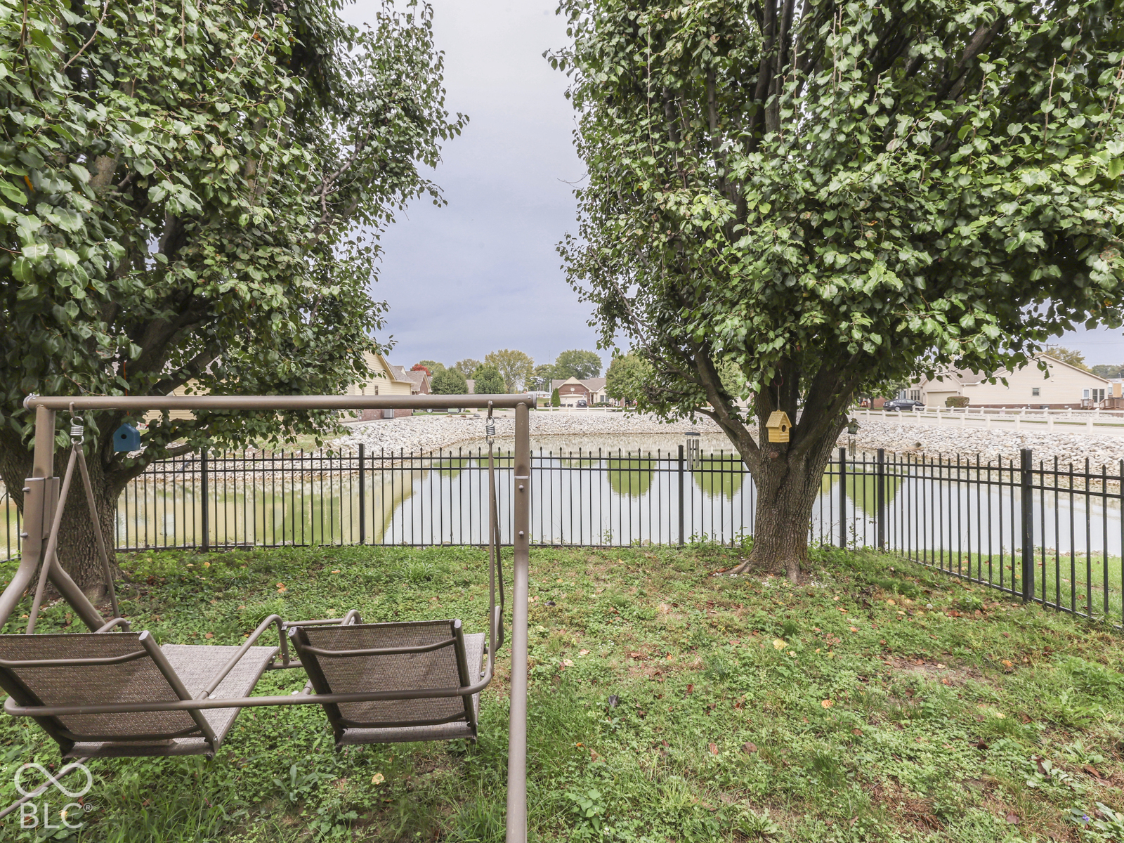 Waterfront backyard featuring mature shade trees, a stone-lined shoreline, and a metal perimeter fence