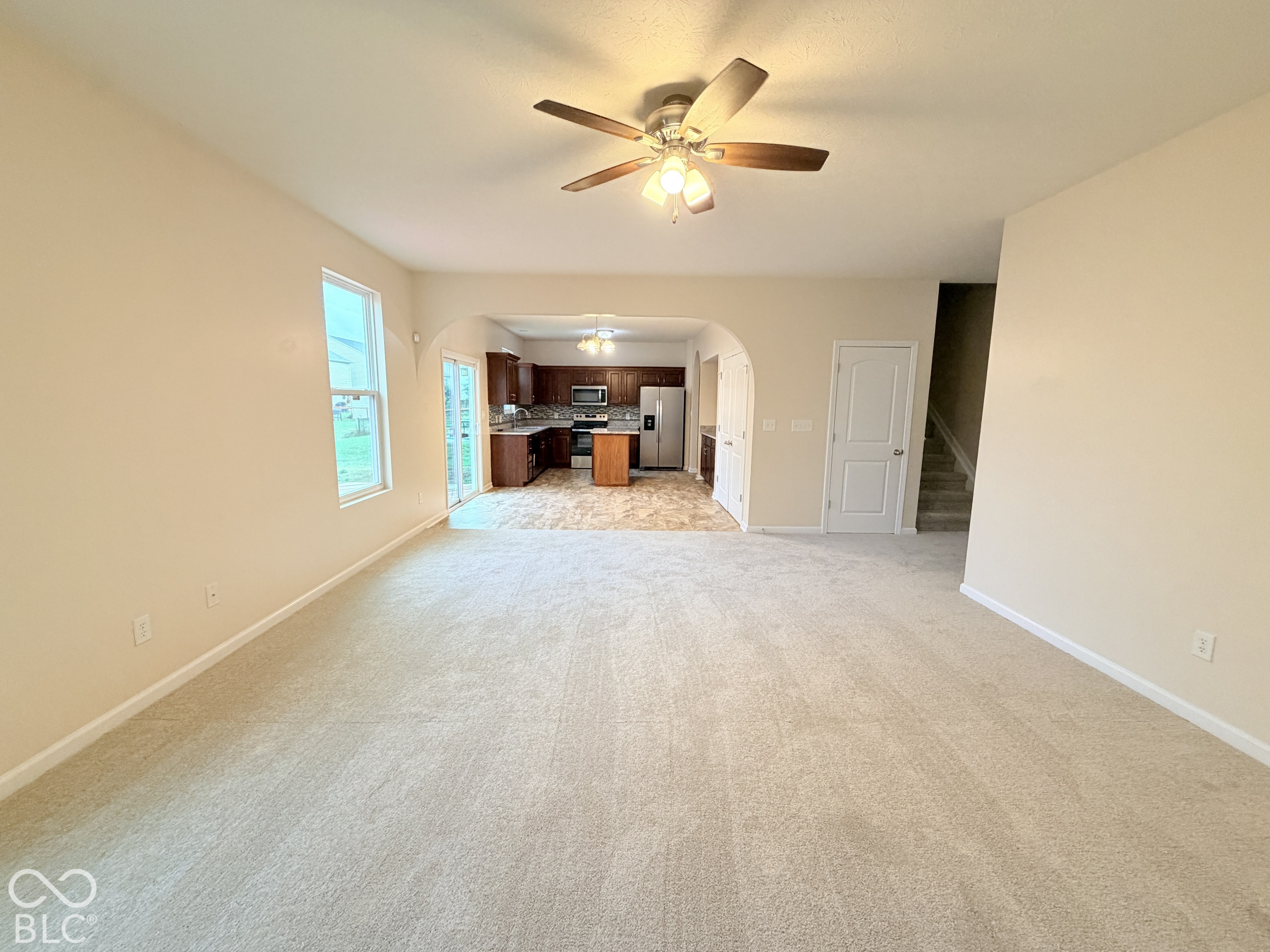Open-concept interior featuring neutral carpeting, an arched entryway, ceiling fan with light fixture, and kitchen cabinetry with stainless steel appliances Open-concept interior featuring neutral carpeting, an arched entryway, ceiling fan with light fixture, and kitchen cabinetry with stainless steel appliances