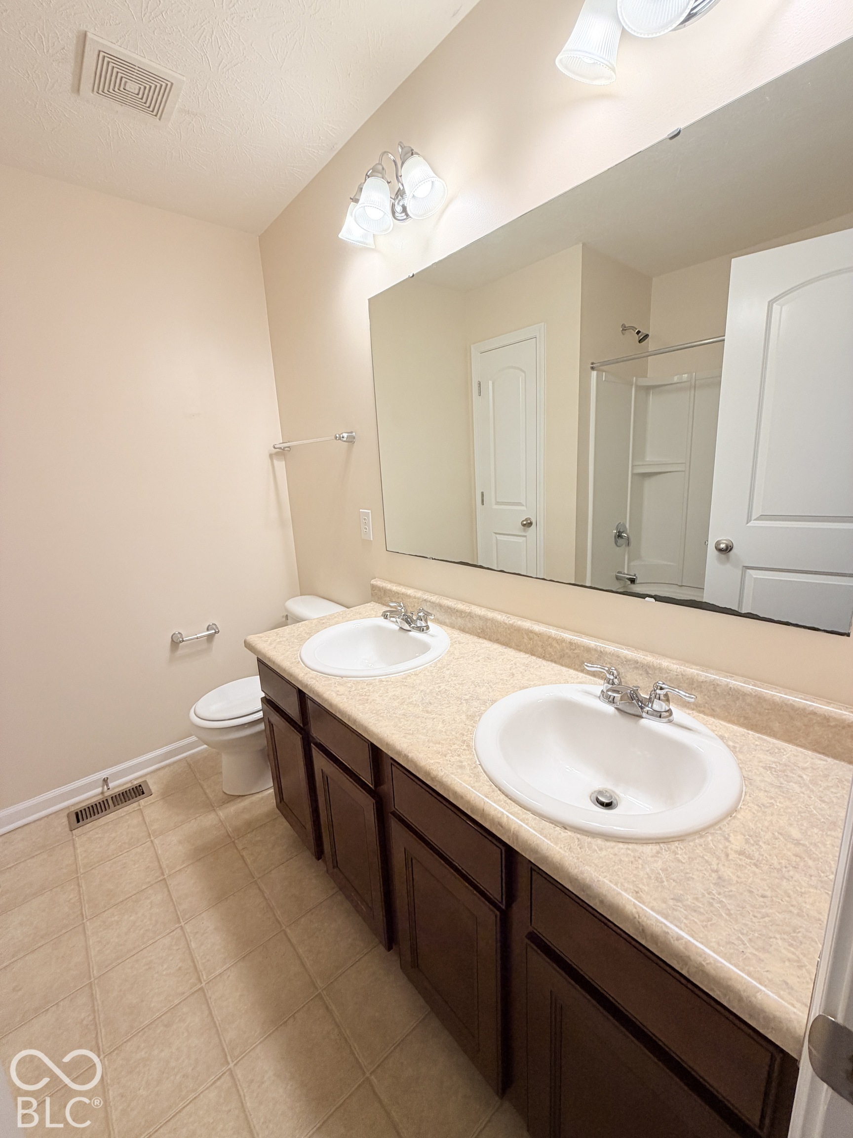 Bathroom featuring a dual-sink vanity with a solid-surface countertop, dark-finish cabinetry, a full-width wall mirror, and a shower-tub combination with an integrated surround