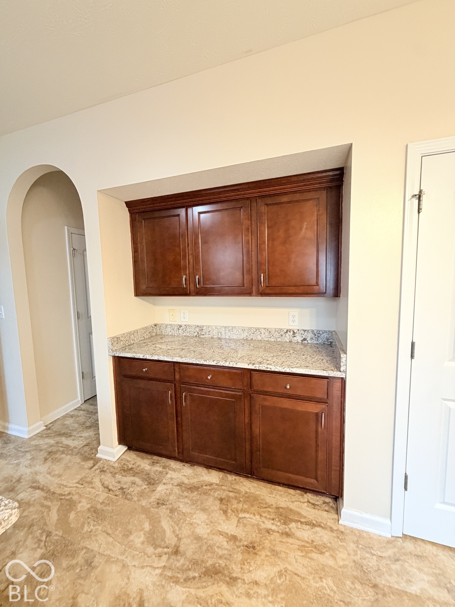 Built-in cabinetry featuring wood-finish lower and upper cabinets, light-toned stone-patterned countertops, and an arched architectural opening