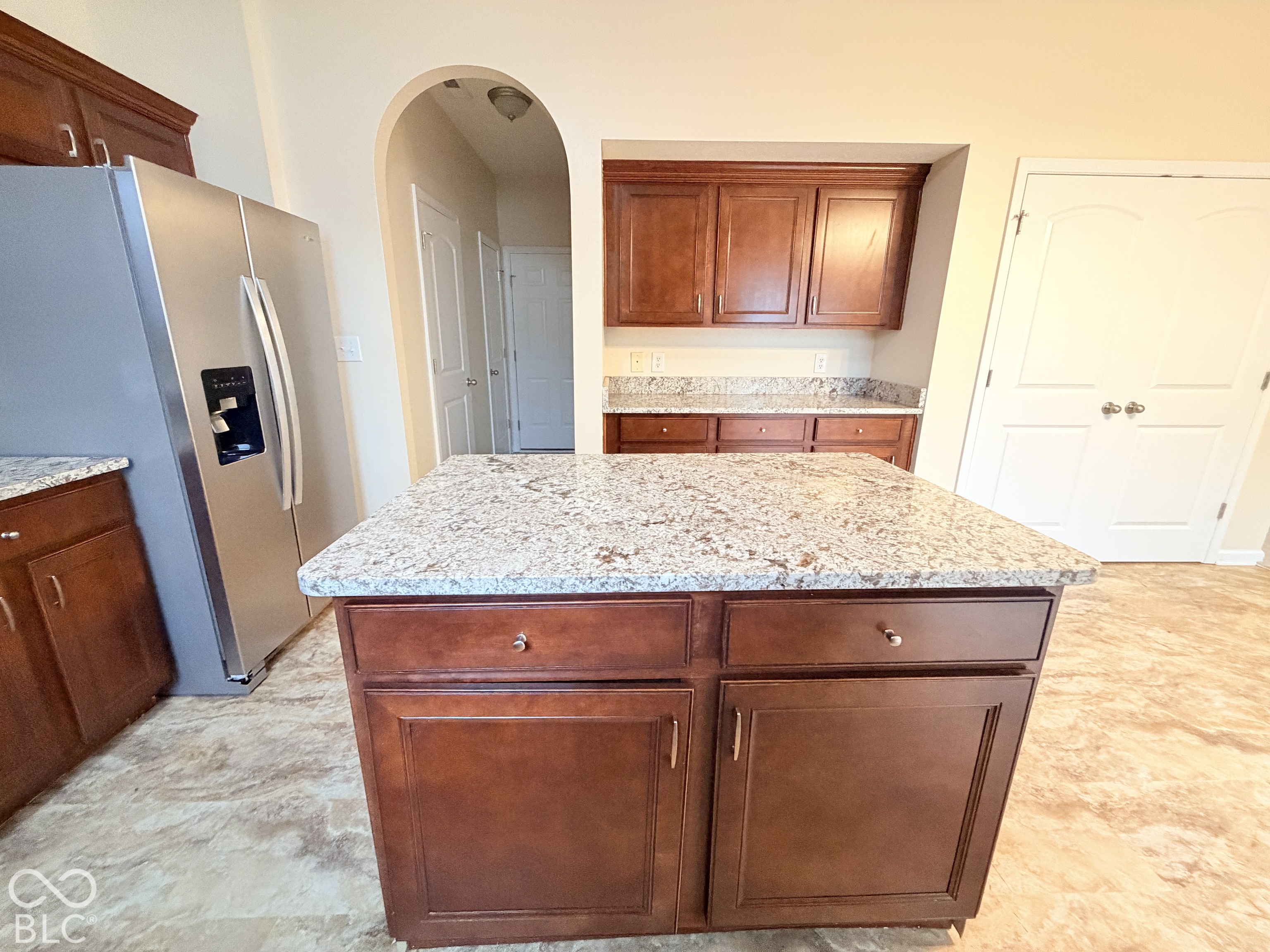 Kitchen featuring a central island with a stone countertop, stained wood cabinetry, stainless steel refrigerator, and arched doorway