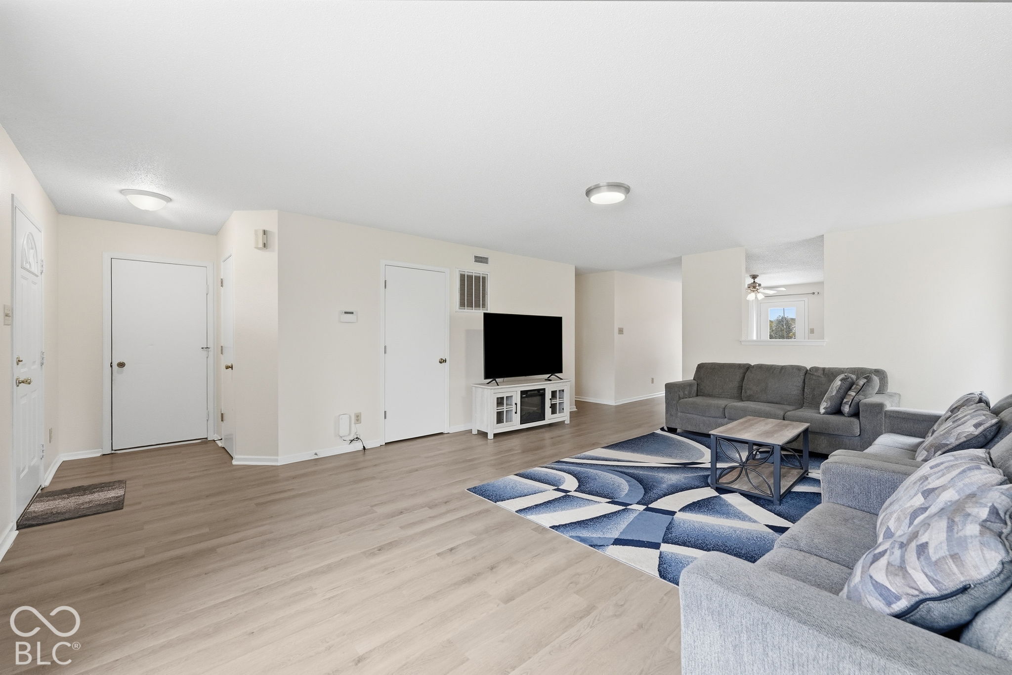 Open-concept living space featuring wood-finish flooring, neutral wall paint, and a pass-through window cutout Open-concept living space featuring wood-finish flooring, neutral wall paint, and a pass-through window cutout