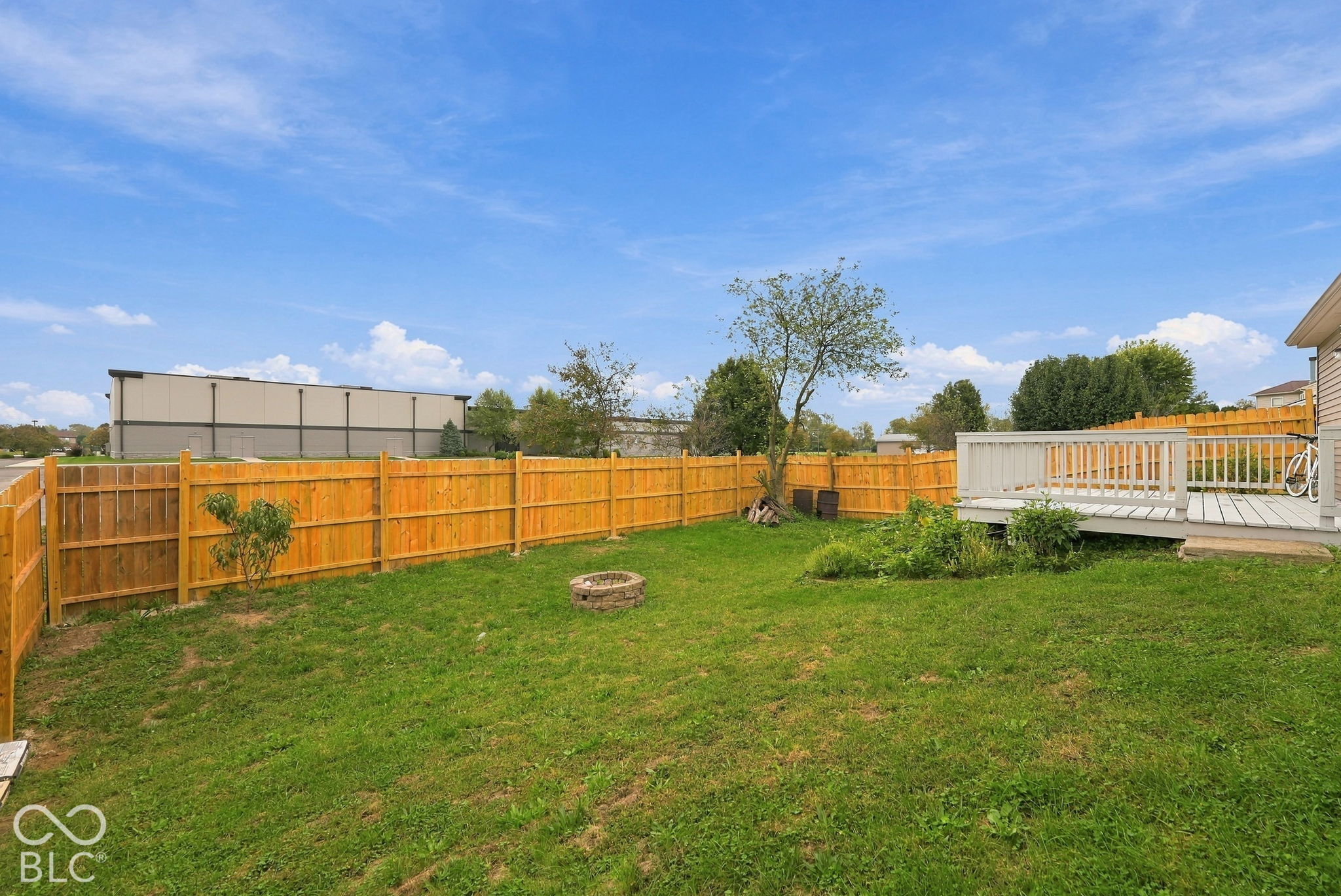 Fenced backyard featuring a raised wooden deck, stone-bordered fire pit, and mature trees
