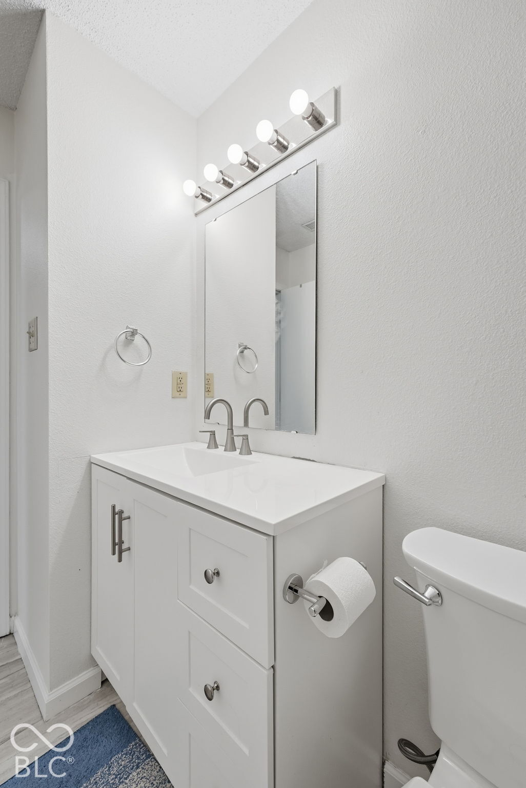 White vanity with shaker-style cabinetry, solid-surface countertop, integrated rectangular sink, wall-mounted multi-bulb light fixture, and polished metal hardware
