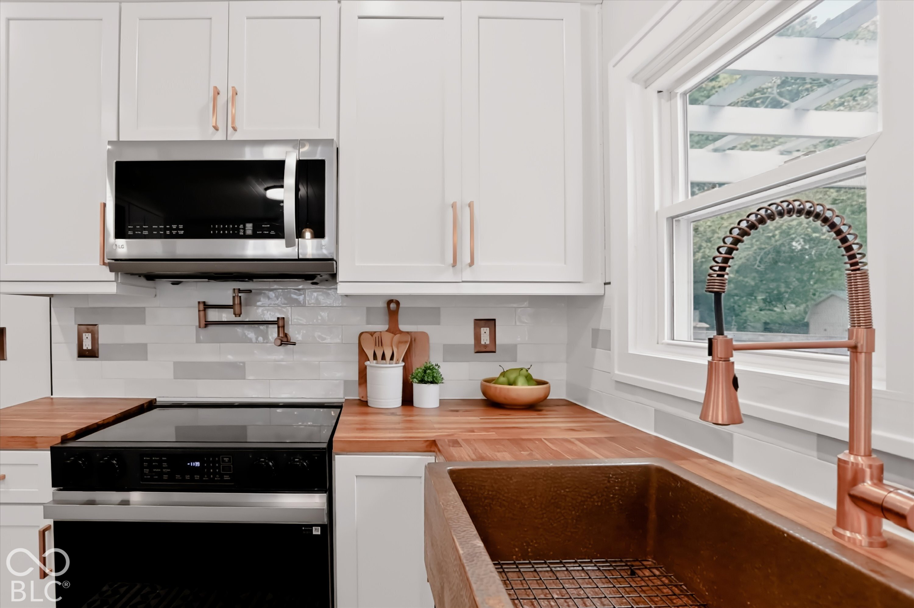 White cabinetry with copper hardware, wood countertops, stainless steel microwave, metal apron-front sink, and industrial-style copper finish faucet