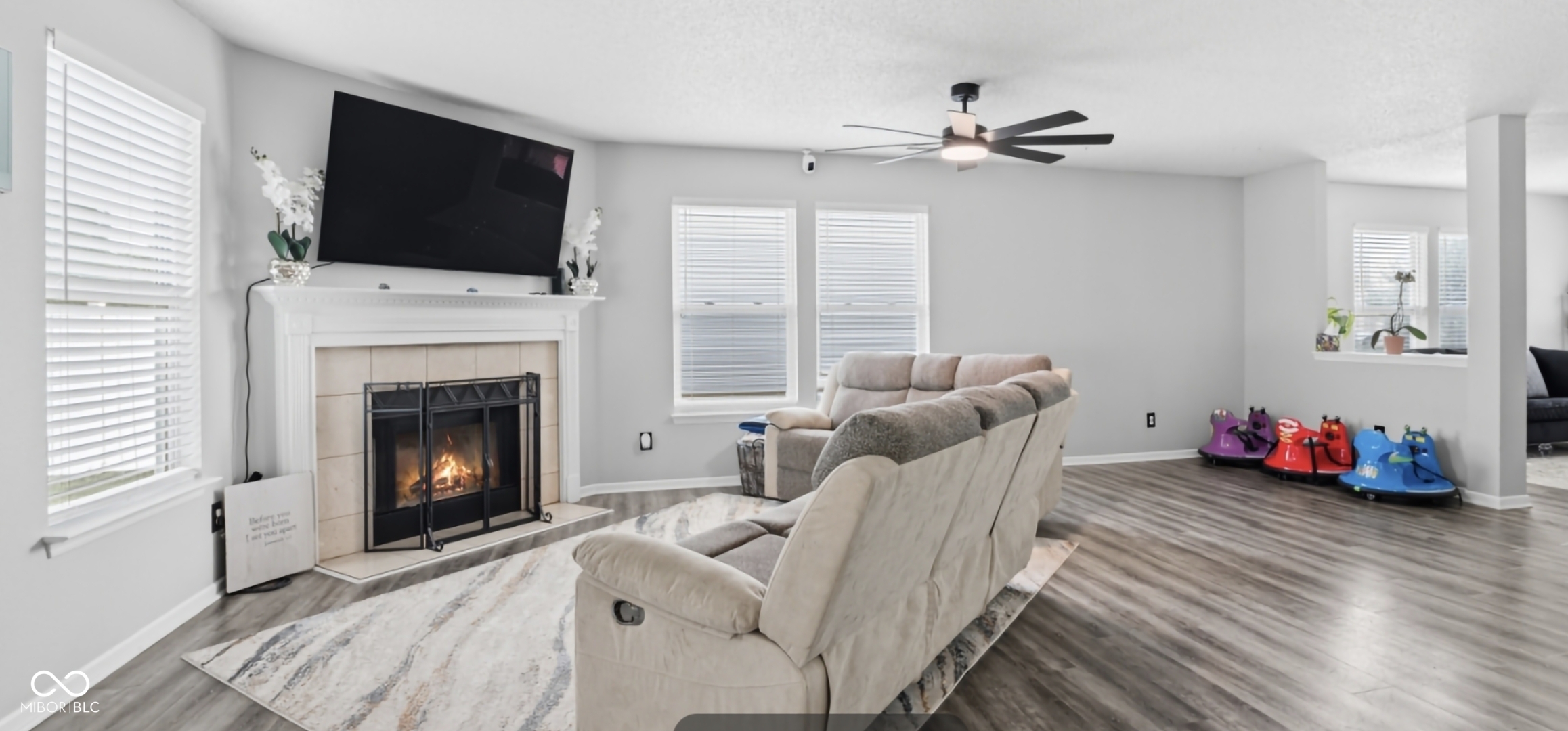 Living area featuring wood-finish flooring, a white mantel fireplace with a tile surround, multiple windows with blinds, and a ceiling fan with integrated lighting