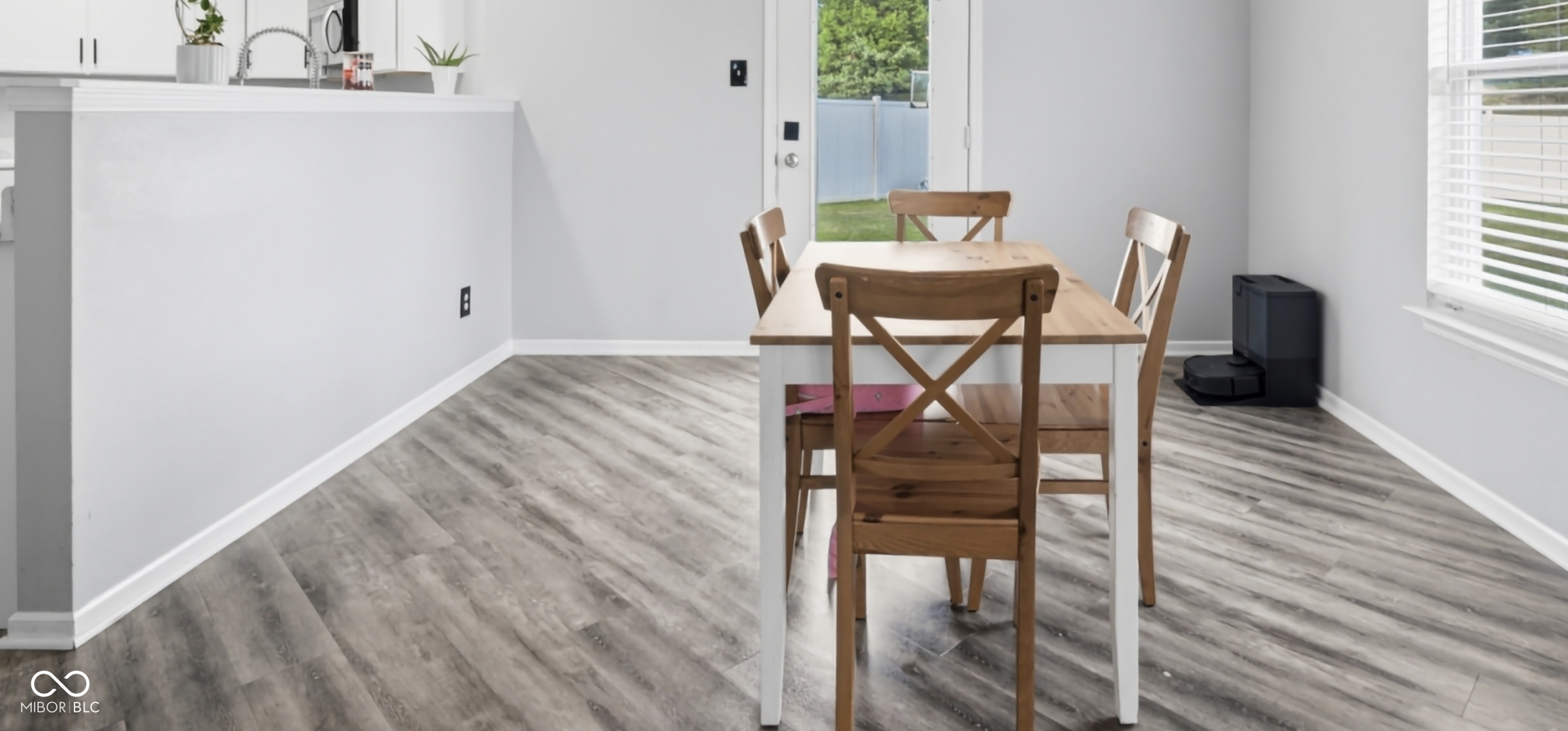 Open-concept dining area featuring wood-finish flooring throughout, a neutral color palette, and abundant natural light from a large window with blinds