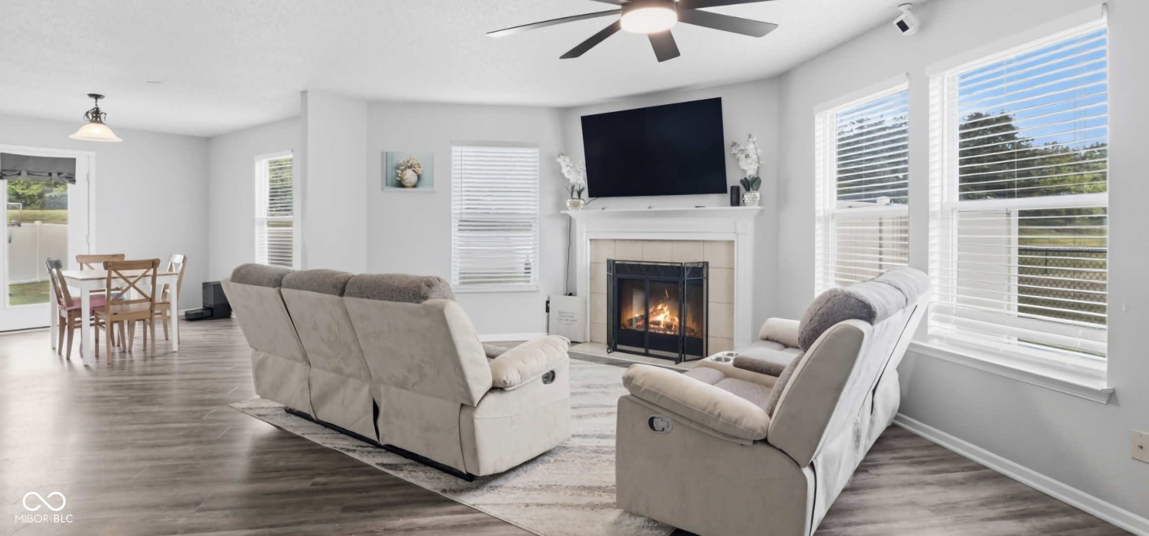 Living area featuring a fireplace with a mantel, ceiling fan with integrated lighting, wood-finish flooring, multiple windows, and a glass-paneled door