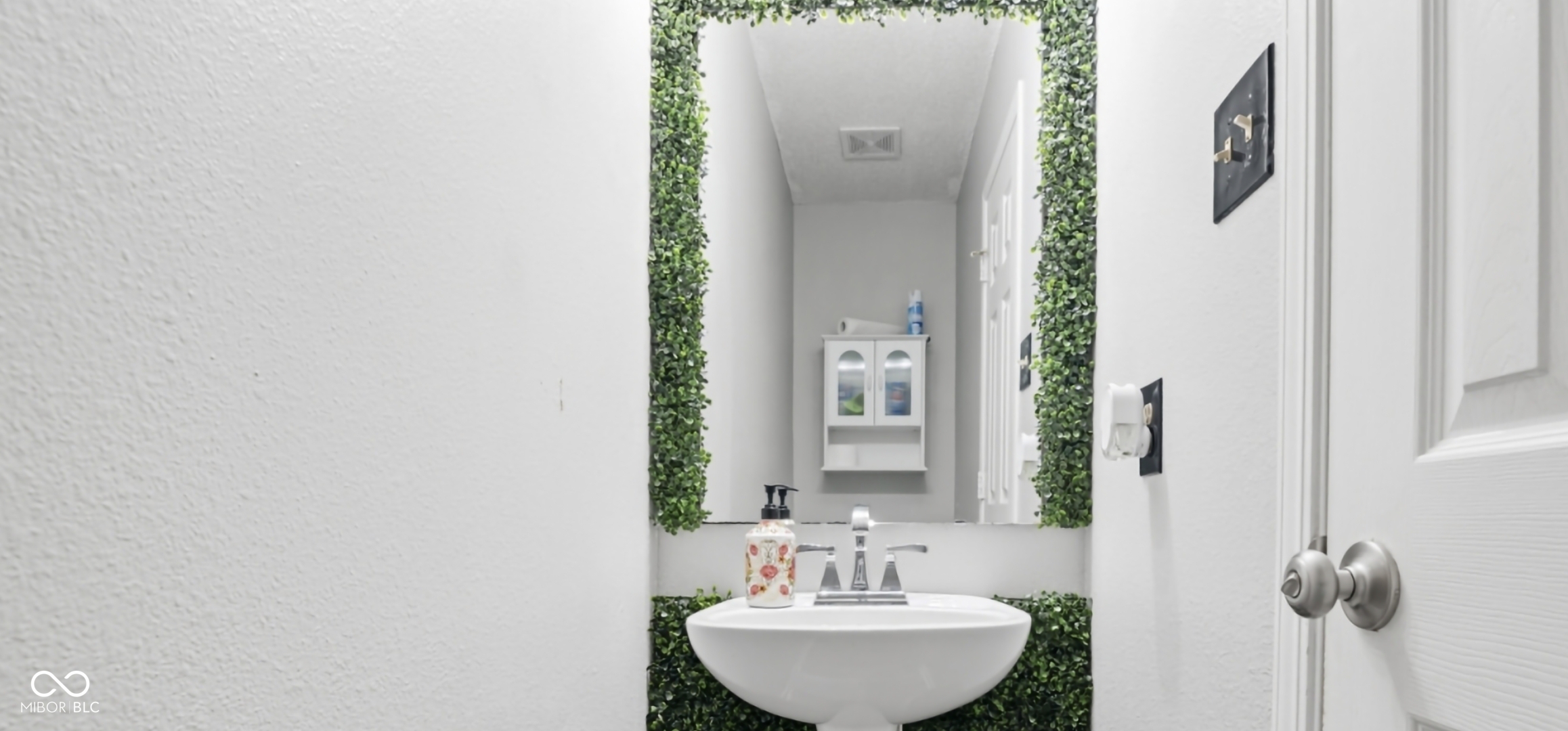 Pedestal sink with a chrome faucet, set against a mirror framed by artificial greenery
