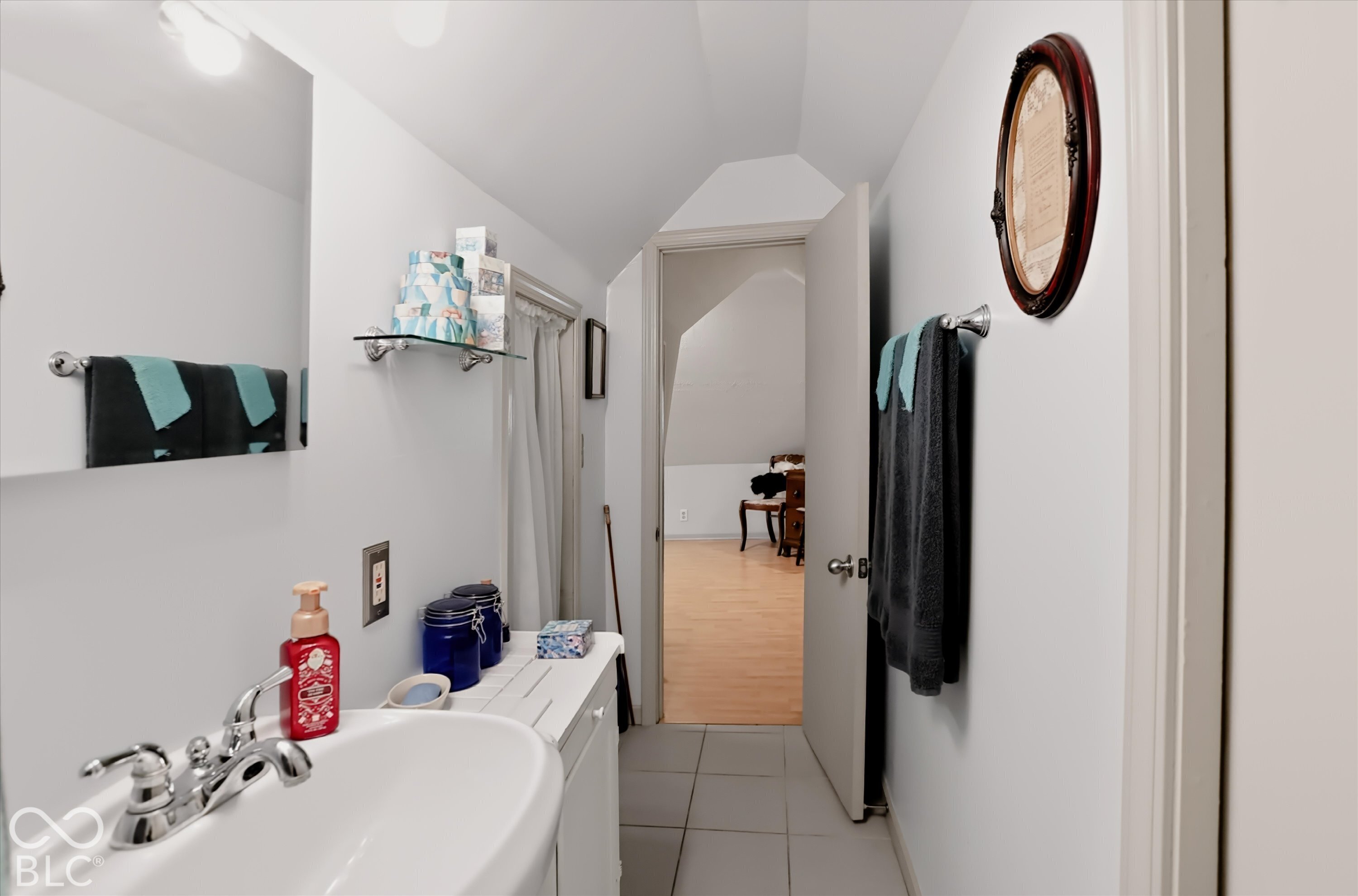 Bathroom featuring a wall-mounted rectangular mirror, vanity with a porcelain sink, polished chrome faucet, glass shelving, and neutral tile flooring