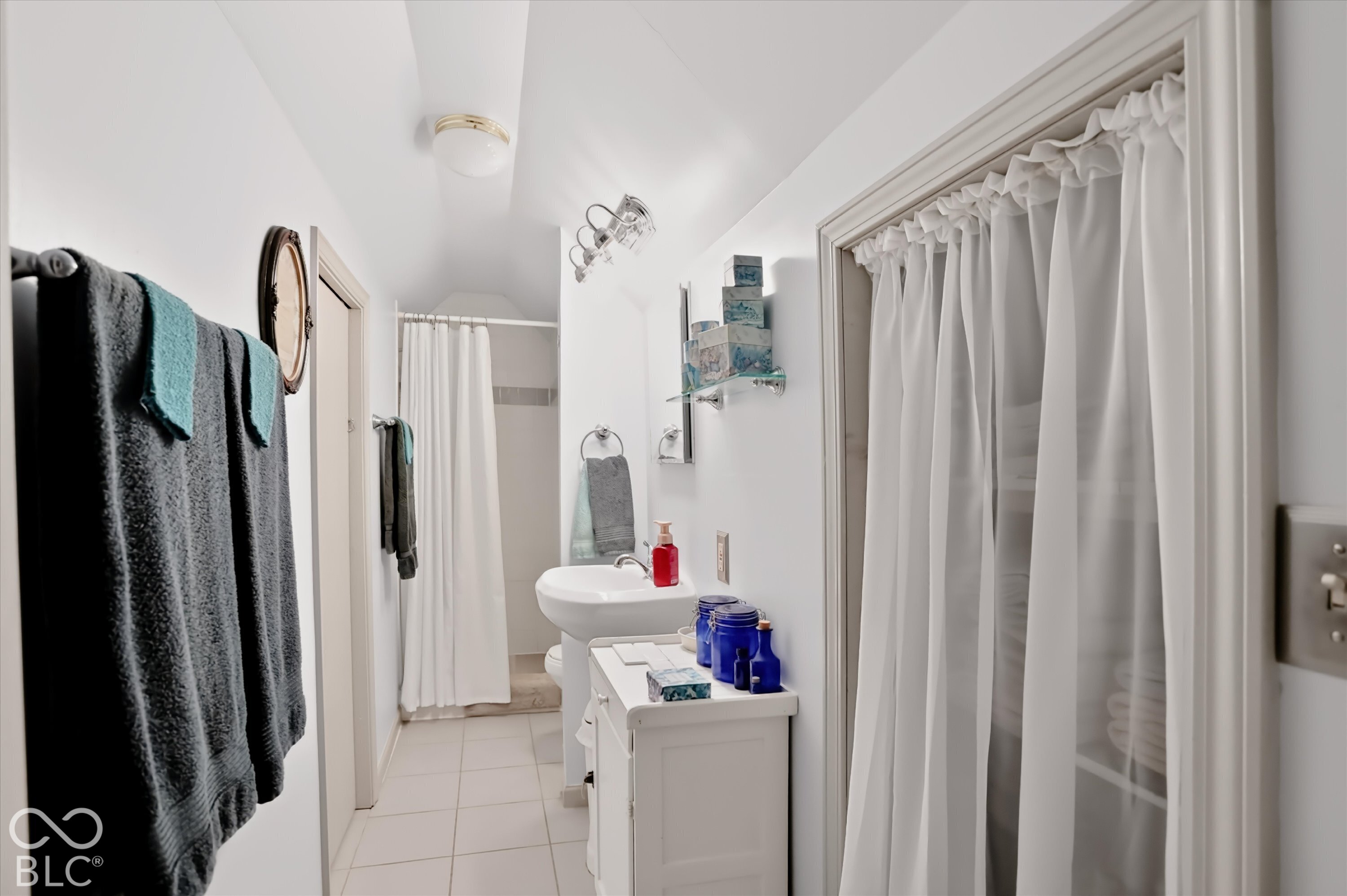 Bathroom featuring a pedestal sink, ceramic tile flooring, built-in shelving with a fabric curtain cover, wall-mounted multi-bulb light fixture, and arched ceiling detail