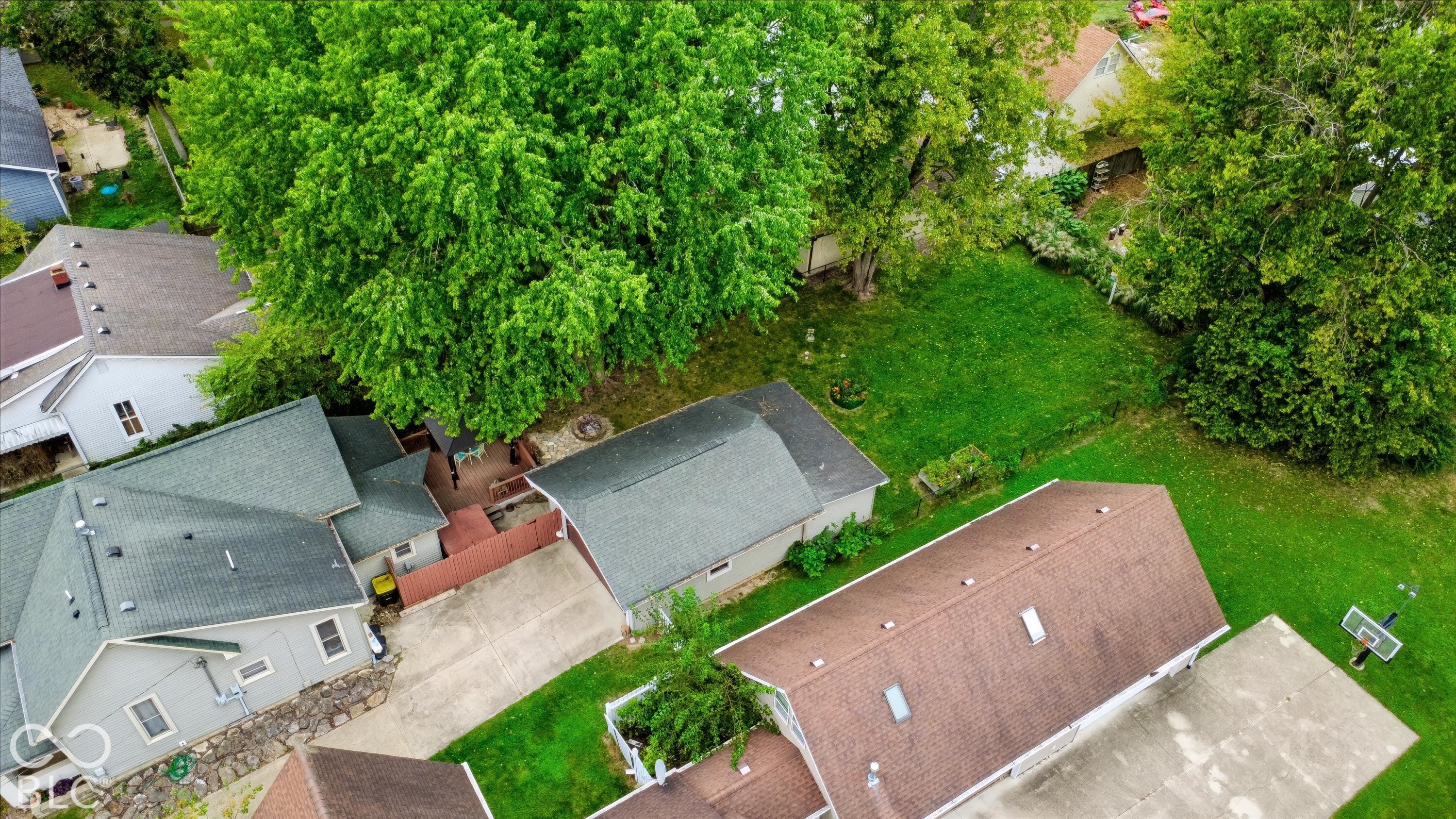 Aerial perspective of a property featuring a concrete driveway, detached garage, hardscaped patio area, and mature trees on a level lot