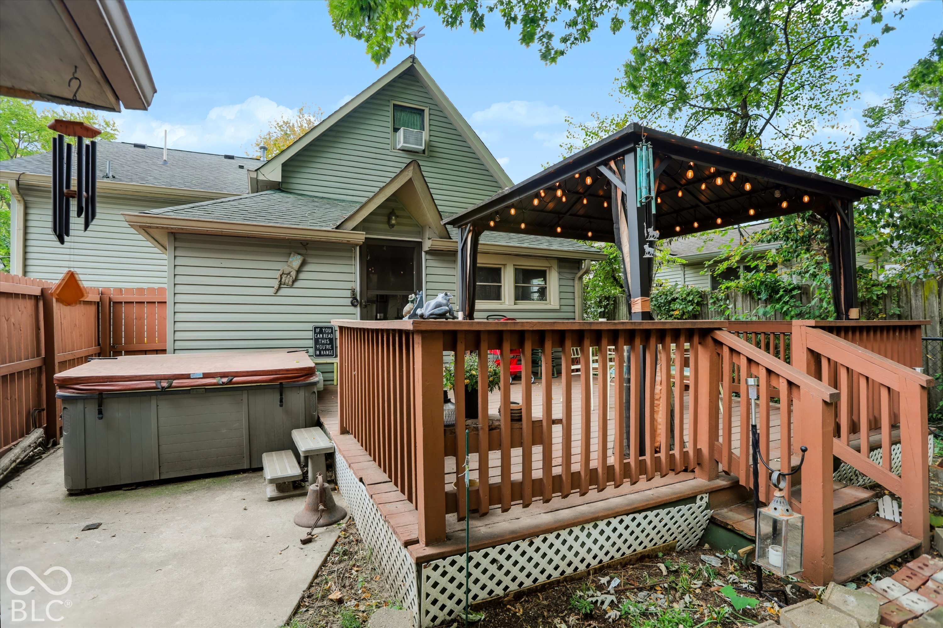 Rear exterior featuring a raised wood deck with a built-in gazebo, wood privacy fencing, concrete hardscaping, and a hot tub