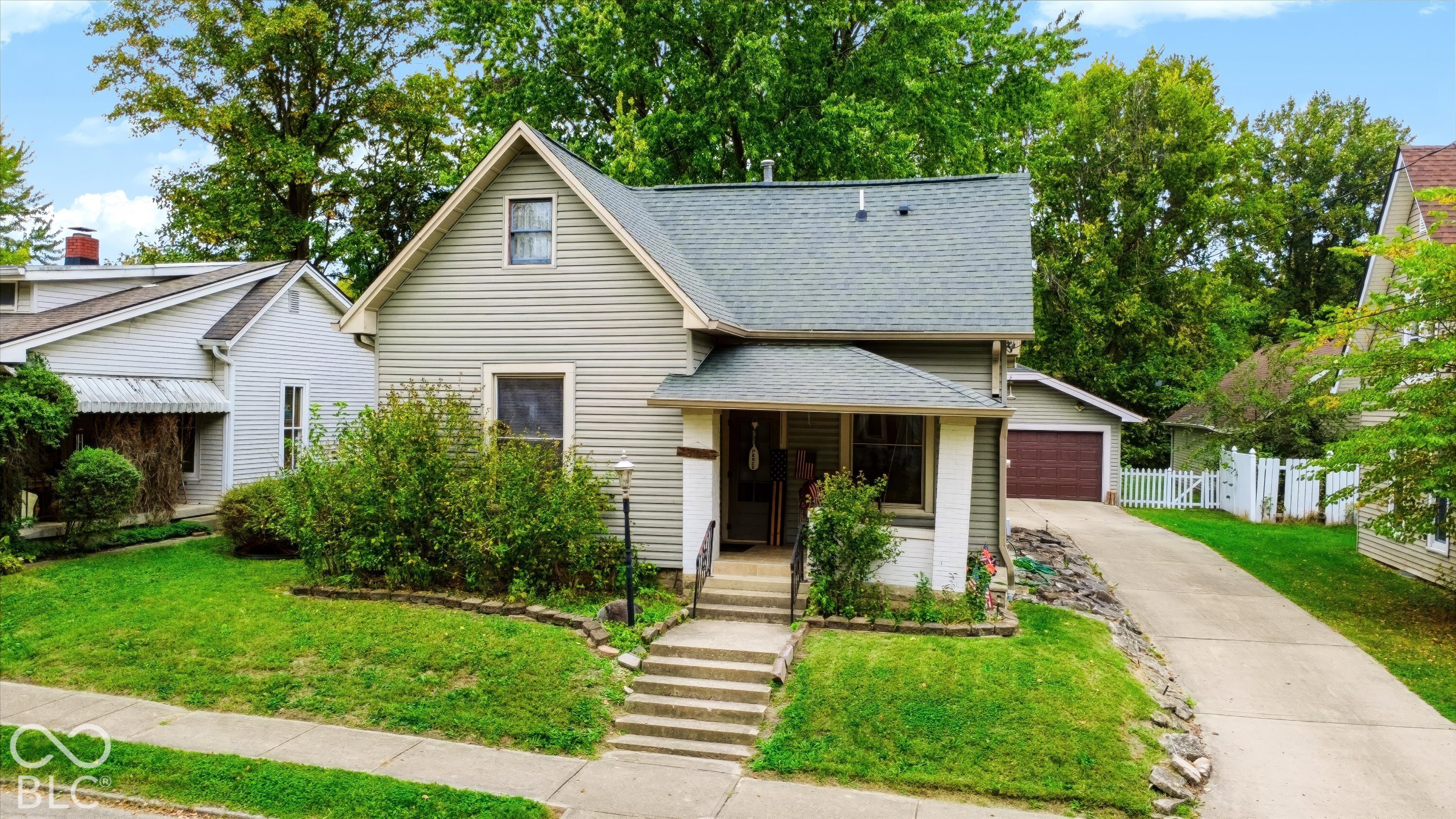 Gable-roofed residence featuring light-toned horizontal siding, a covered front porch with squared columns, concrete steps, a private driveway, and a detached garage
