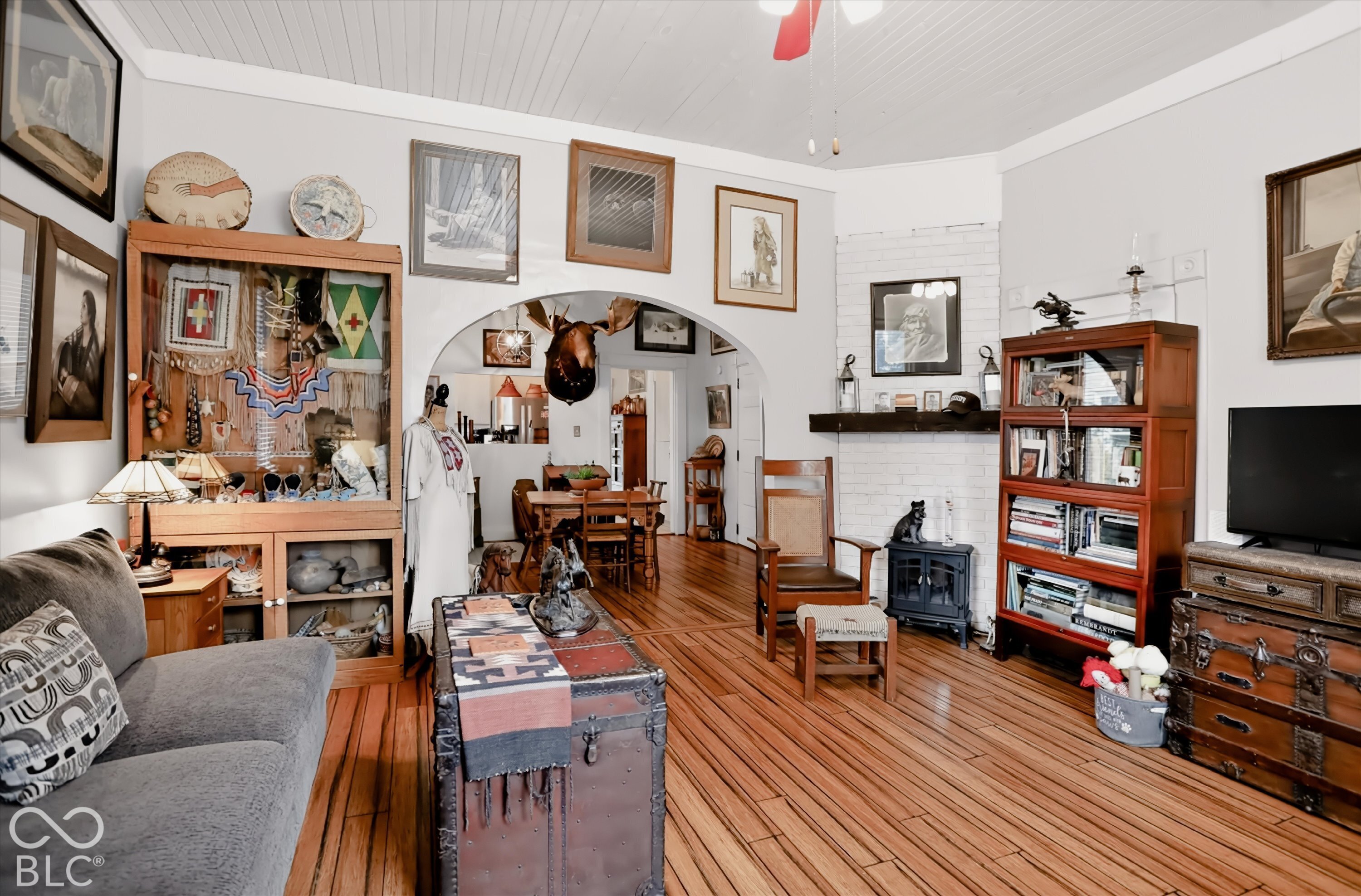 Living area featuring wood-finish flooring, a painted brick fireplace surround, an arched entryway, and beadboard ceiling paneling