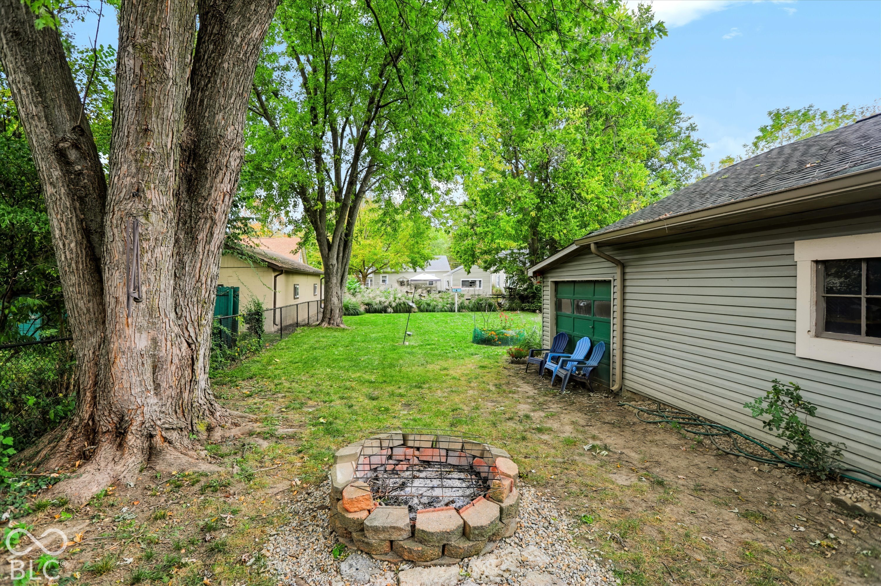 Backyard featuring a stone-lined fire pit, mature trees, chain-link fencing, and a detached garage with horizontal siding