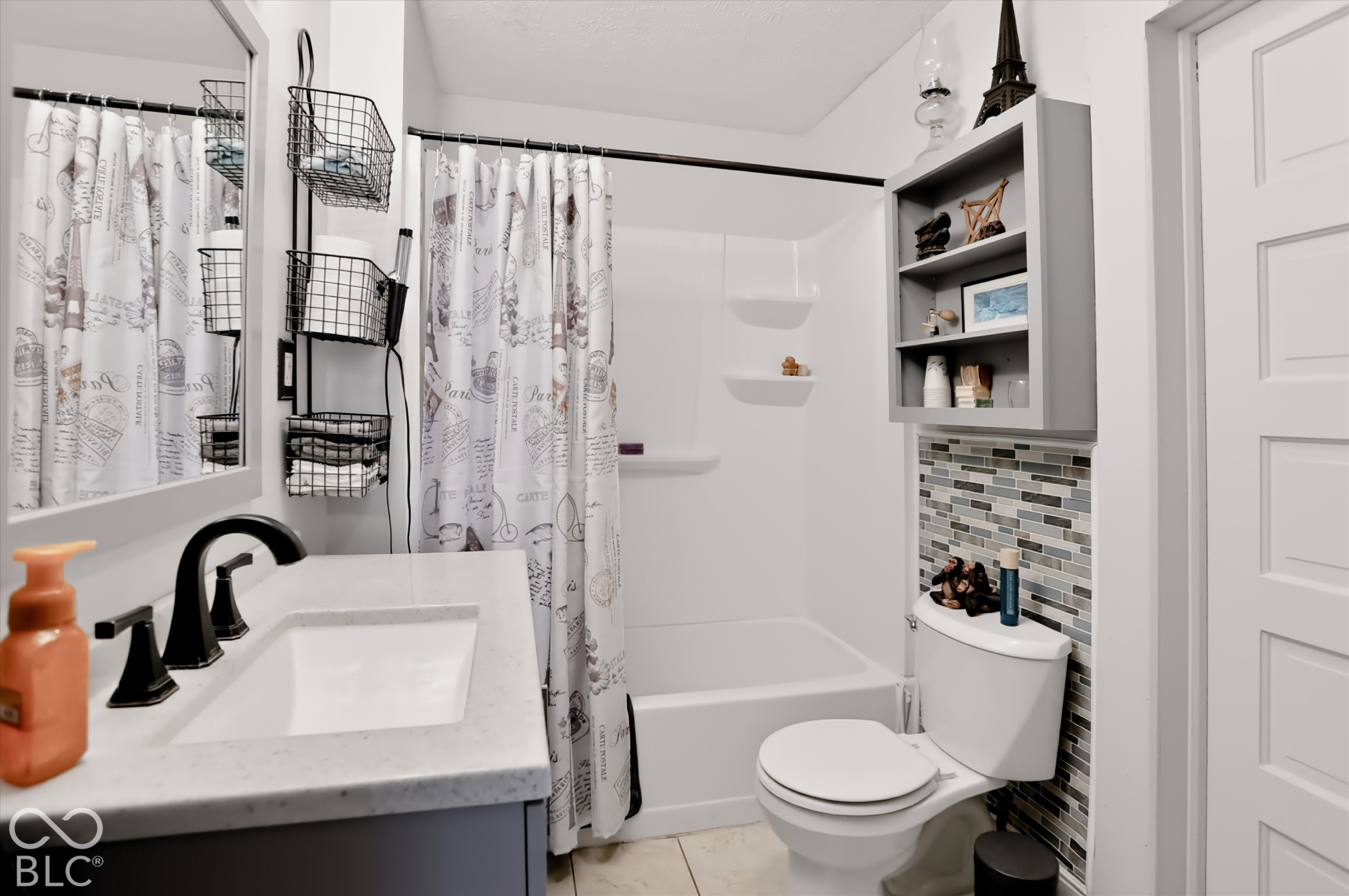 Bathroom featuring a vanity with stone-composite countertop, undermount sink, and matte black faucet, accented by a glass and stone tile backsplash, integrated shower-tub enclosure with built-in shelving, and wall-mounted cabinetry