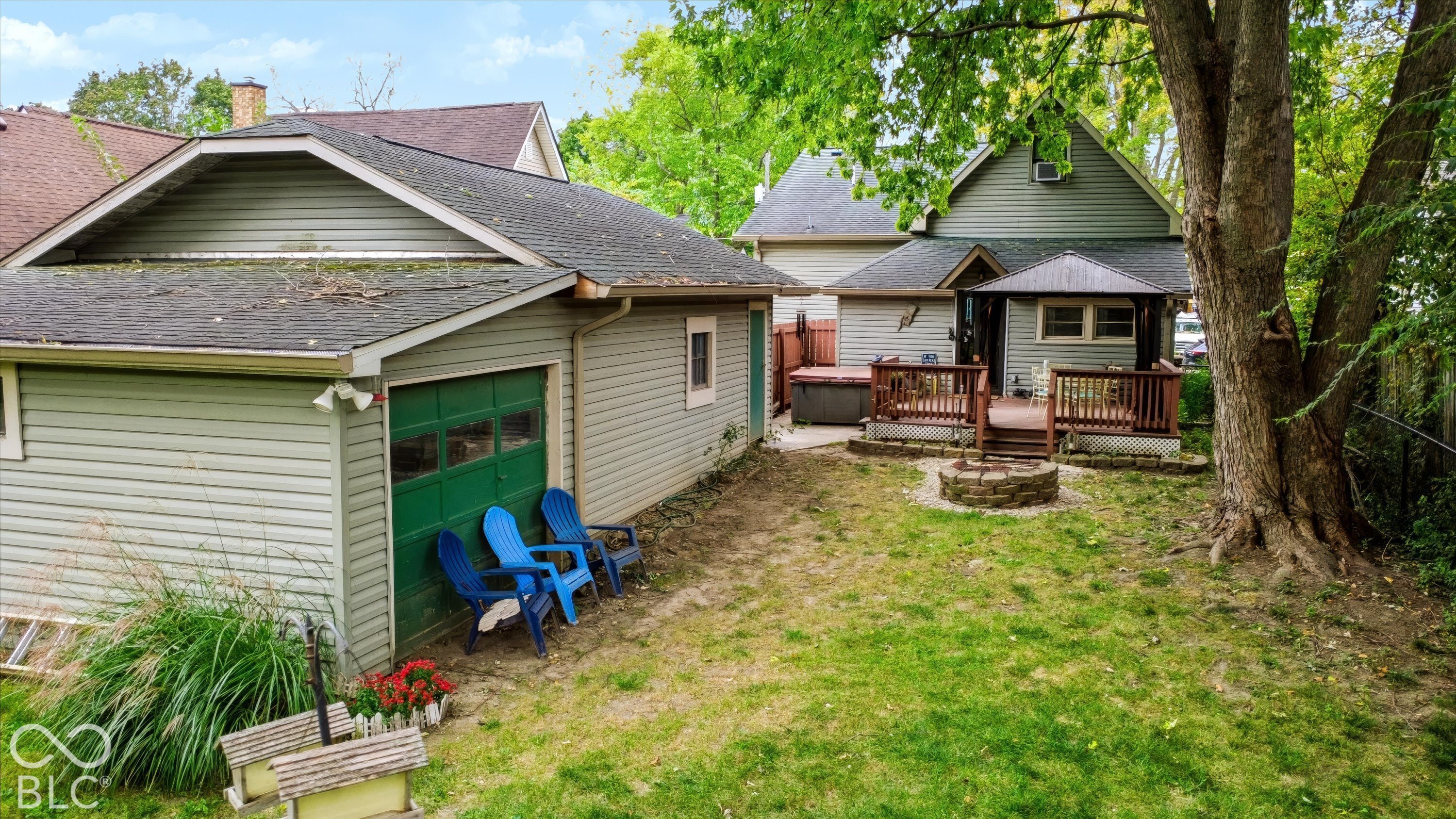 Backyard featuring a detached garage with green paneled door, wood-decked patio, stone fire pit, and mature trees