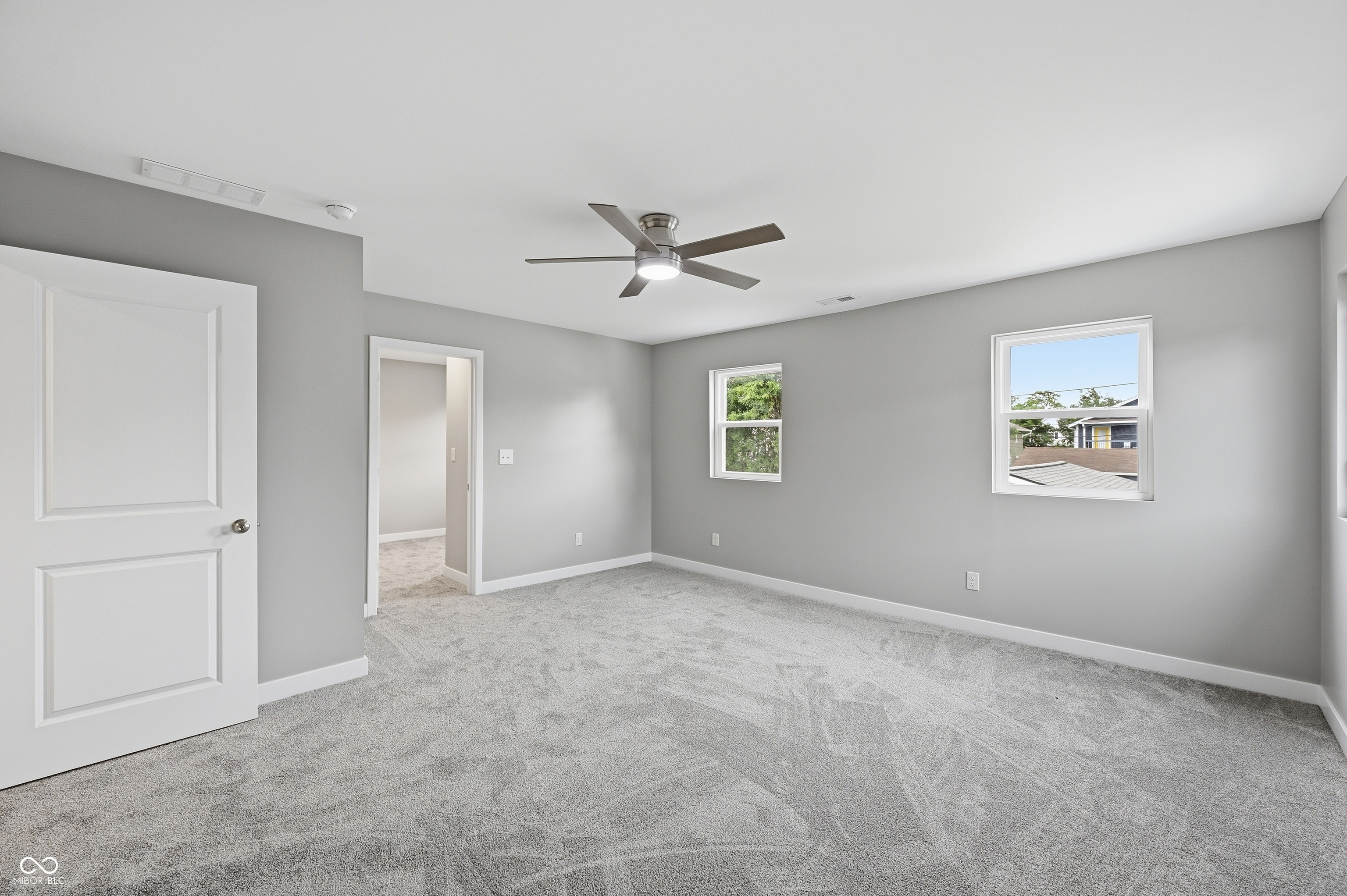 Carpeted room with light gray walls, featuring a modern ceiling fan, two windows providing natural illumination, and a white paneled door