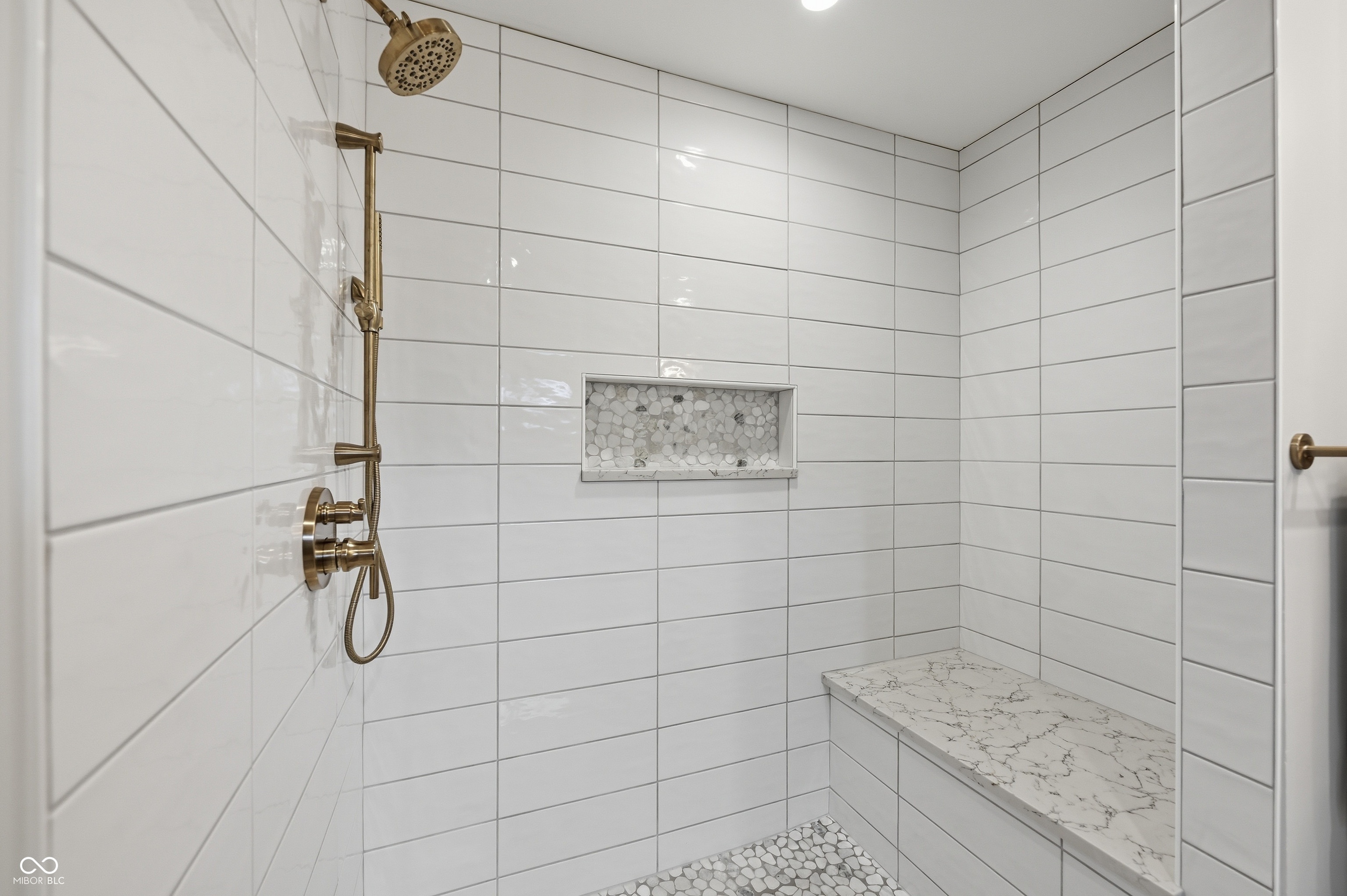 Shower with white rectangular tile walls, a built-in bench with a stone-finish top, a recessed niche with pebble-style tile, bronze-finish shower fixtures, and pebble-style tile flooring