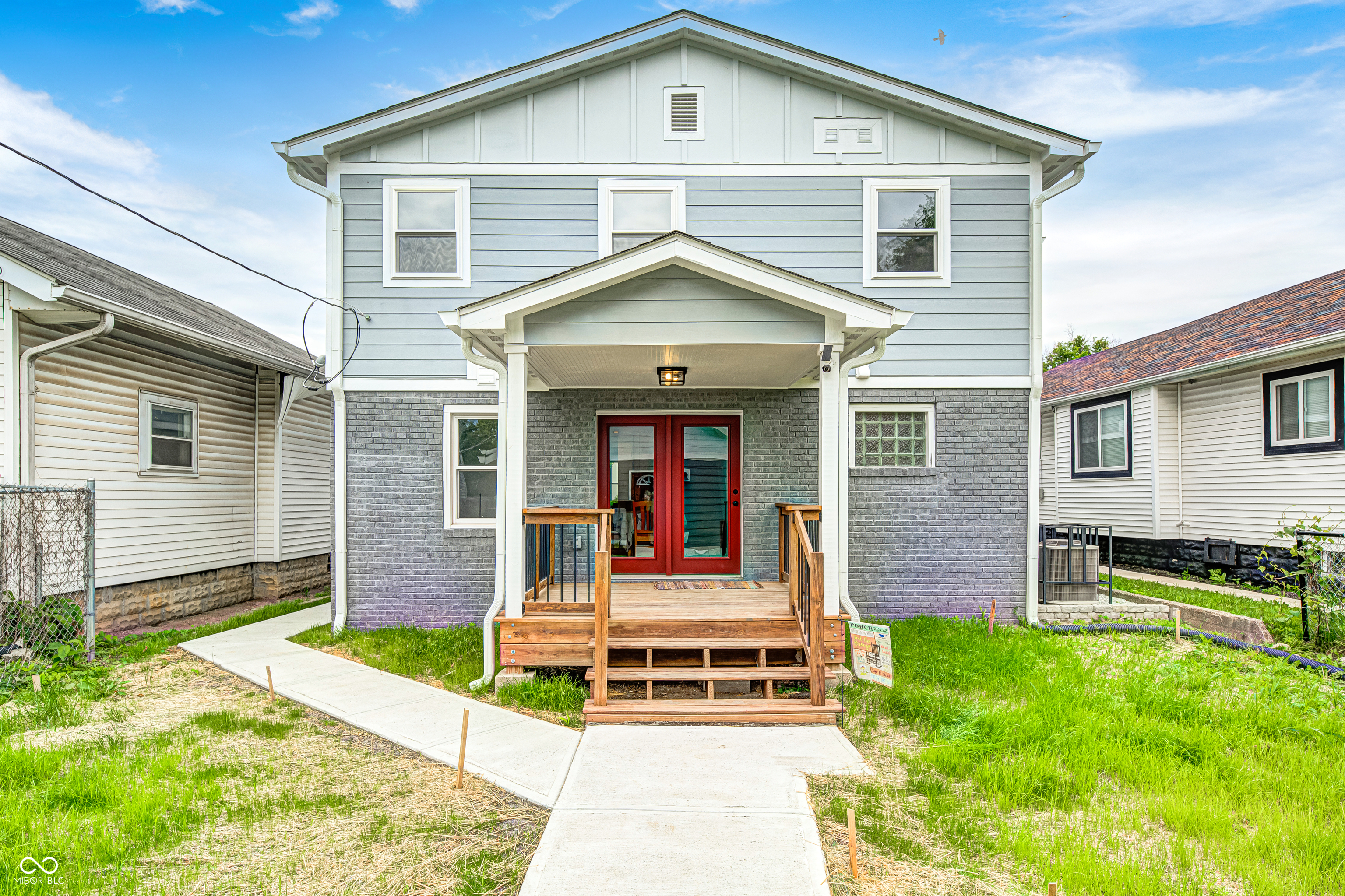 Two-story exterior featuring light blue siding with grey brickwork, a covered porch, wood-tone steps, and a bold red entry door