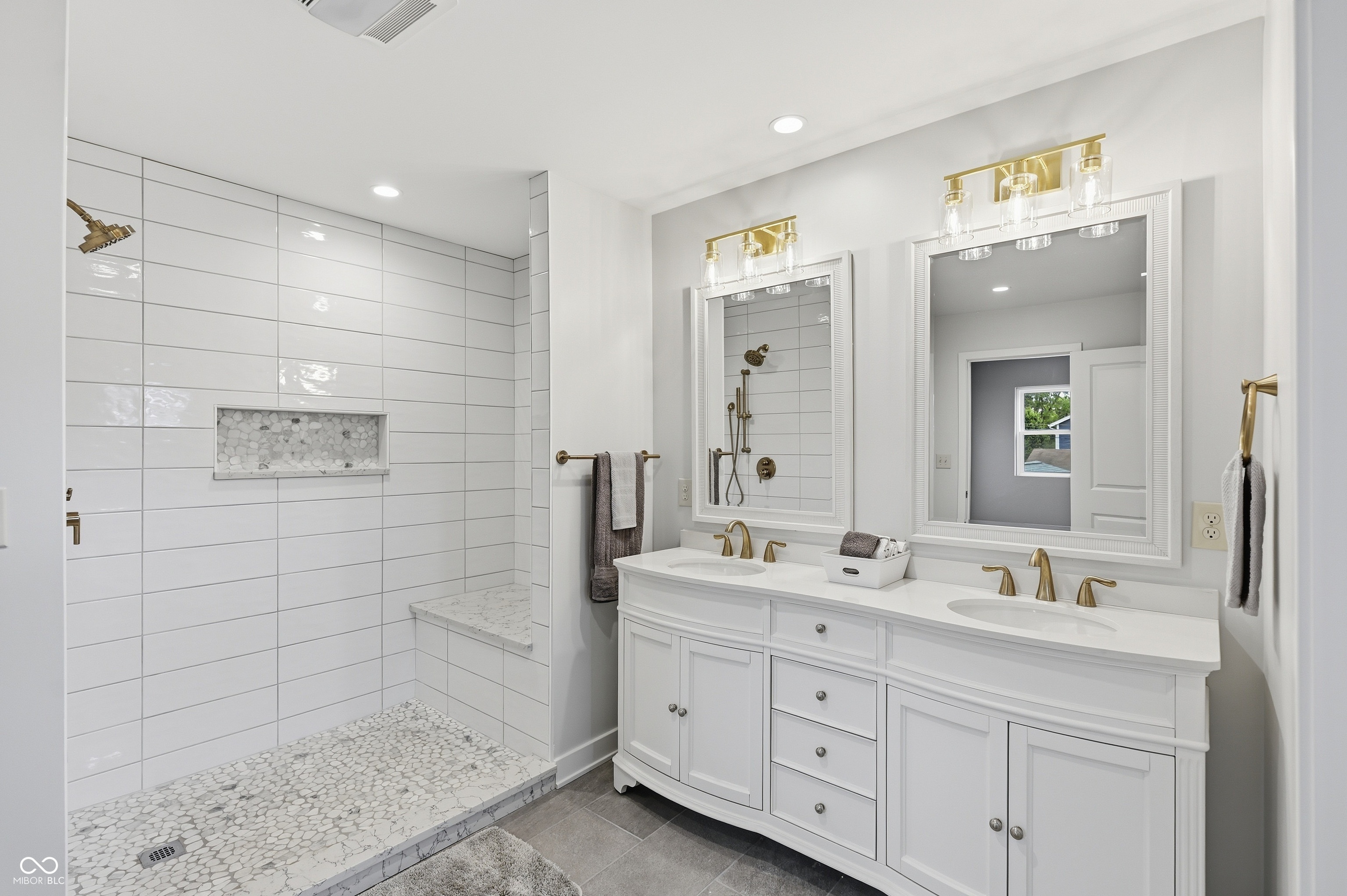 Spacious bathroom featuring a double vanity with white cabinetry and undermount sinks, bronze-finish fixtures, dual framed mirrors with overhead lighting, and a walk-in shower with white subway tile, pebble flooring, and a built-in bench