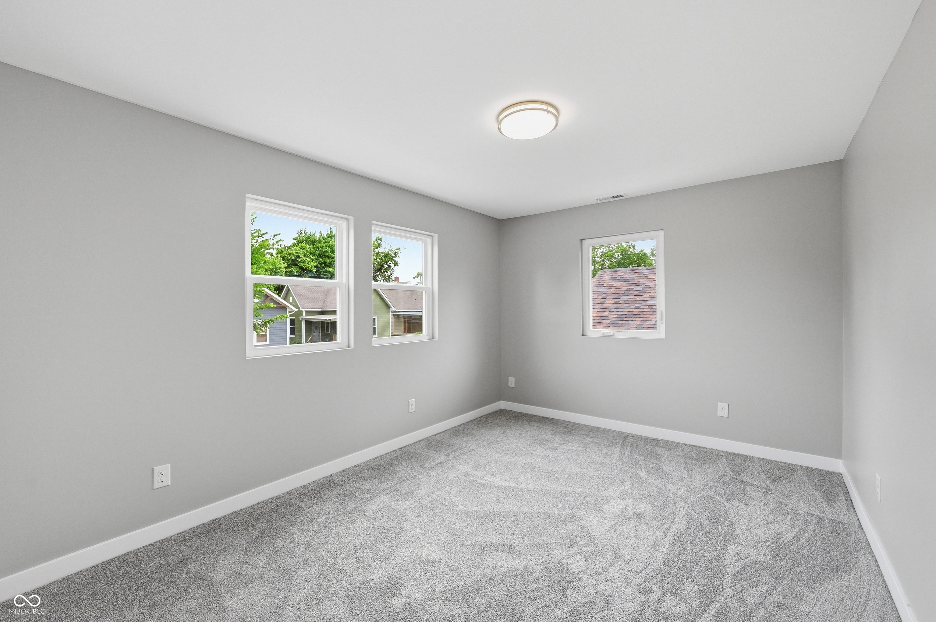 Spacious room featuring light gray walls, plush gray carpeting, white trim, three windows, and a flush-mount ceiling light fixture