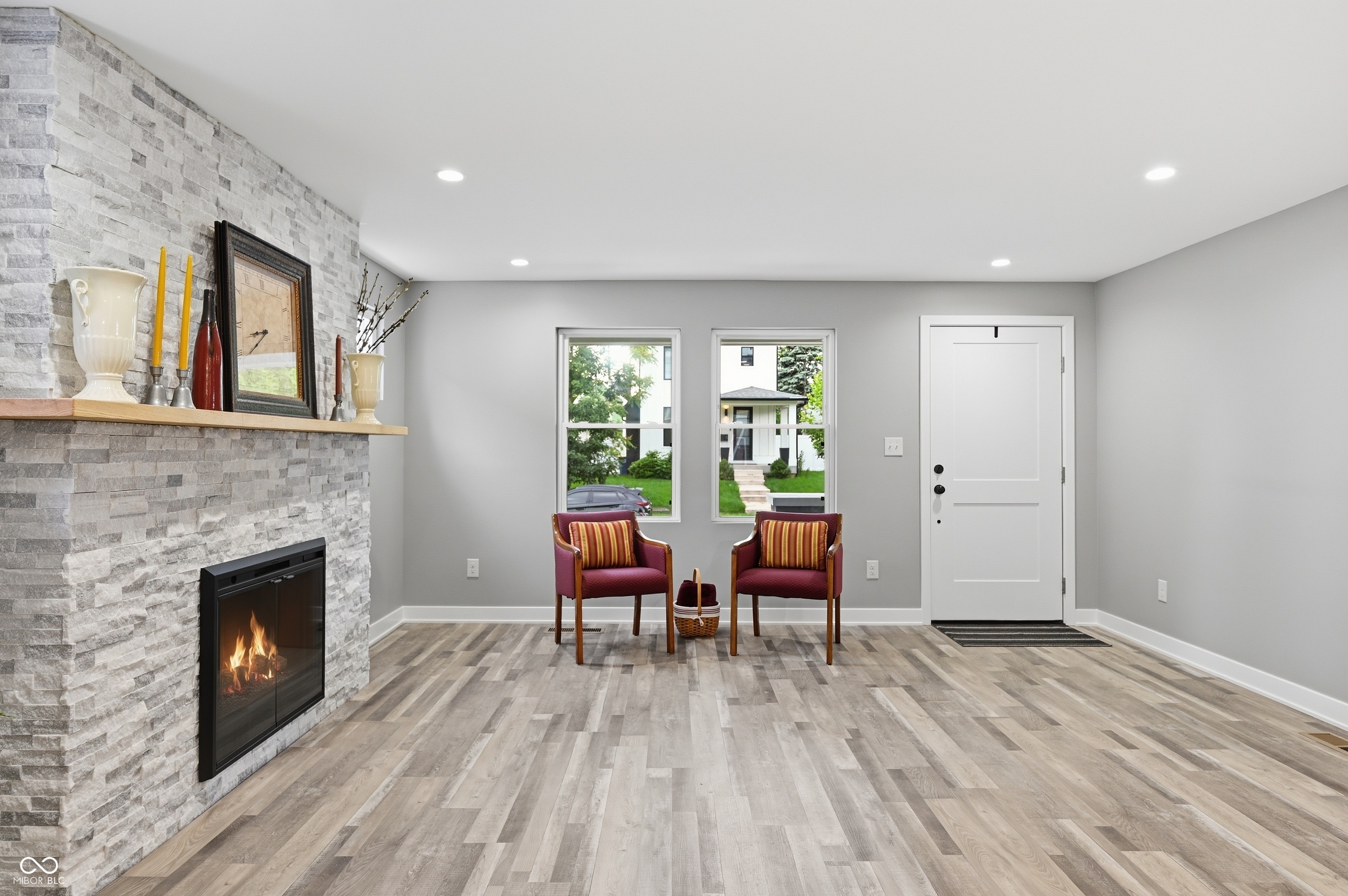 Light-filled living space featuring wood-finish flooring, a stone-clad fireplace with a wood mantel, recessed lighting, and a paneled entry door Light-filled living space featuring wood-finish flooring, a stone-clad fireplace with a wood mantel, recessed lighting, and a paneled entry door