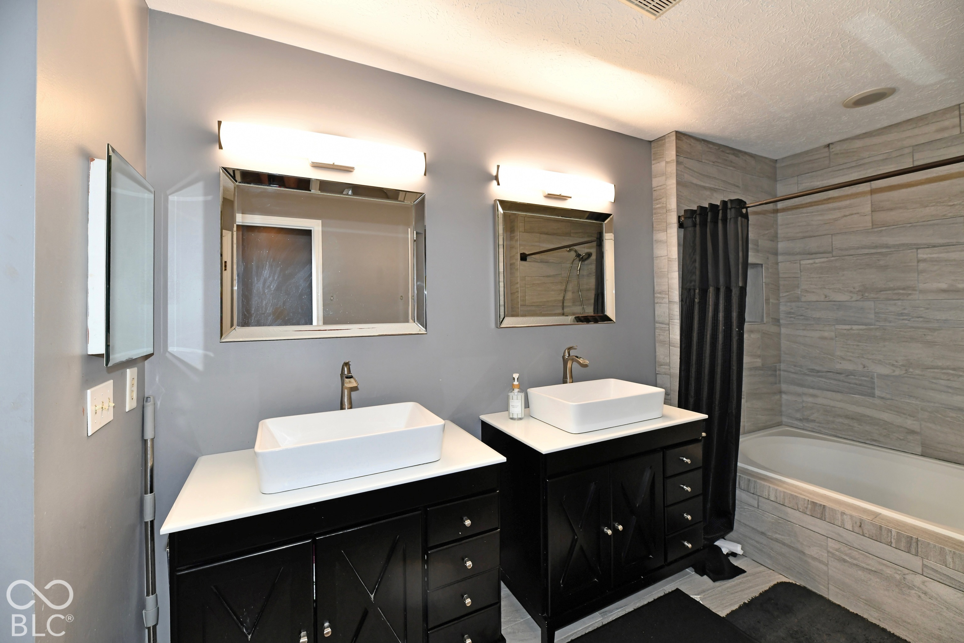 Dual vanity featuring white vessel sinks, quartz countertops, black cabinetry with X-pattern detailing, horizontal wall-mounted lighting, and a tile-surround bathtub