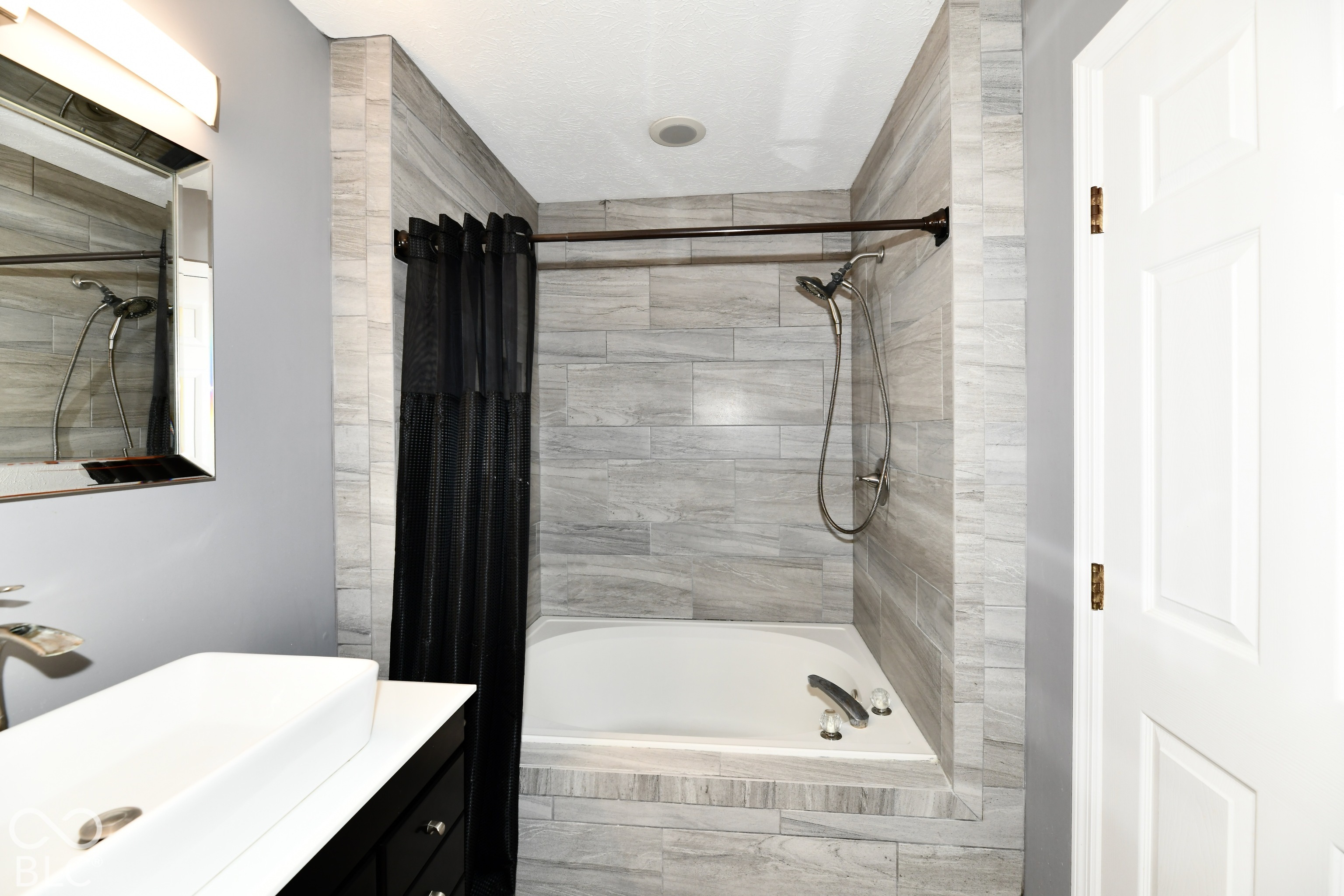 Recessed soaking tub featuring neutral-toned stone-finish tile surround, handheld shower fixture, rectangular vessel sink atop a dark vanity, and wall-mounted vanity lighting
